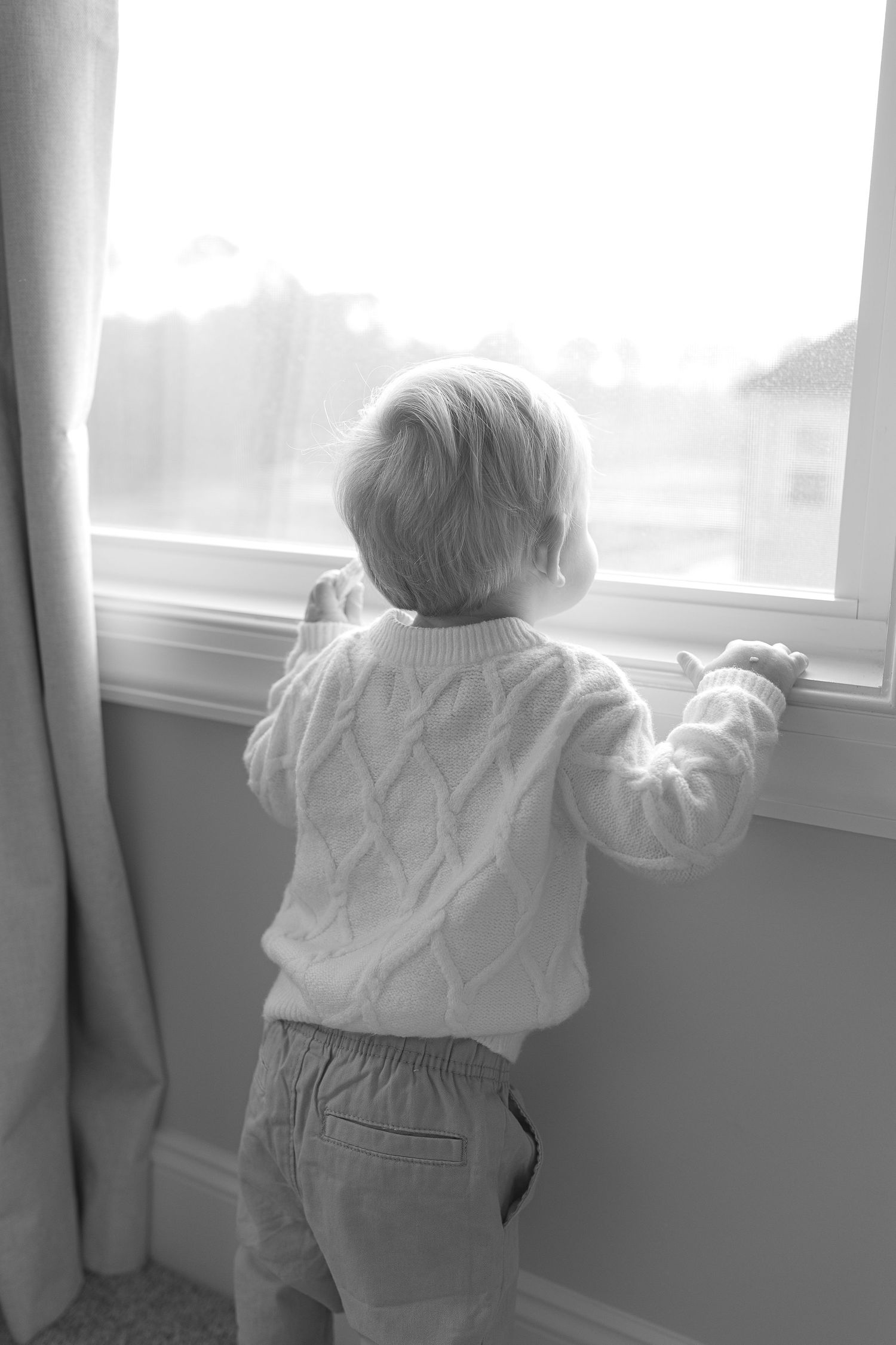 Black and white photo of a toddler in a cable knit sweater looking out a window.  | Karina Thomson Photography Raleigh NC