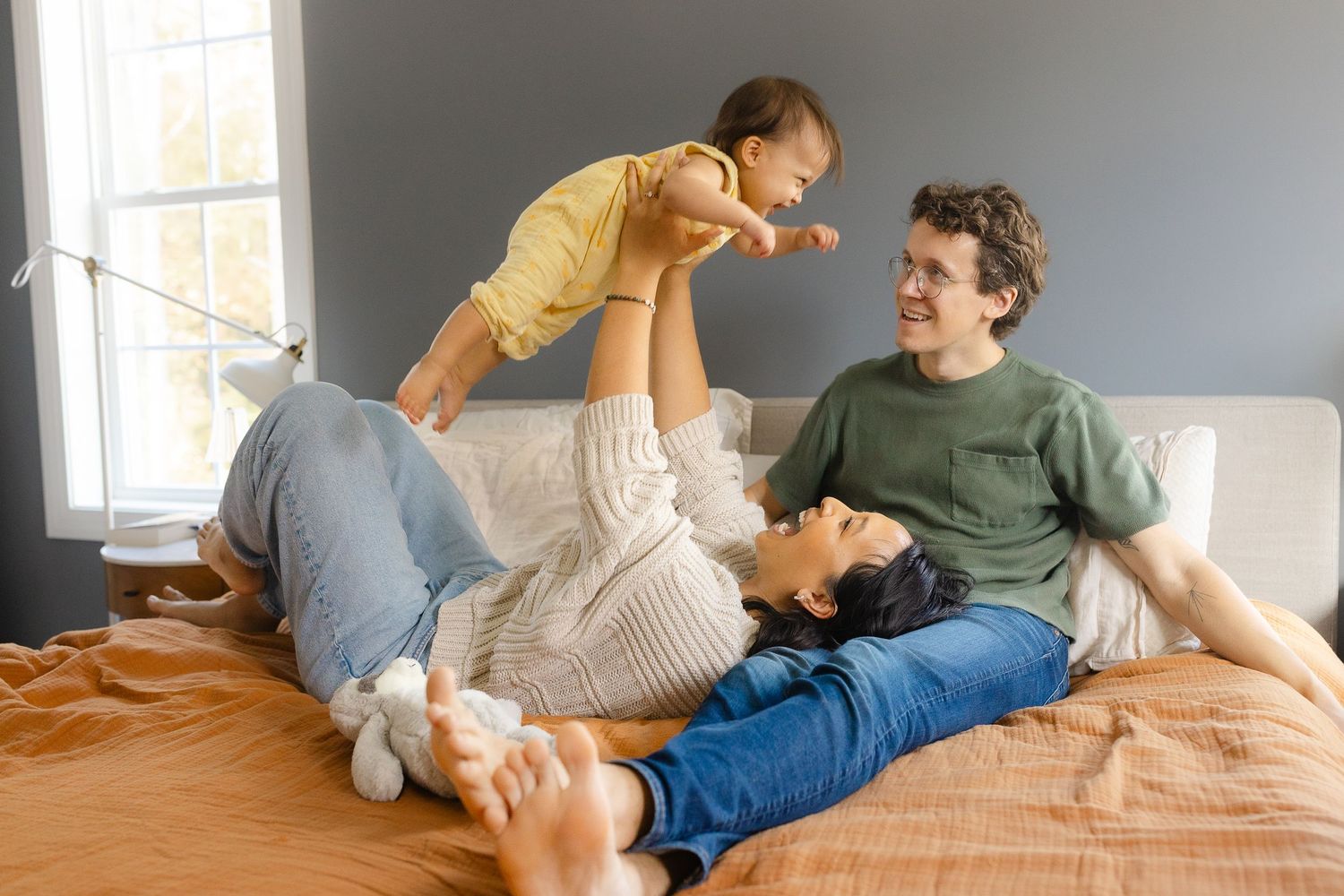 Playful family moment on bed as mother lifts laughing baby in yellow onesie while father watches joyfully.  | Karina Thomson Photography Ral