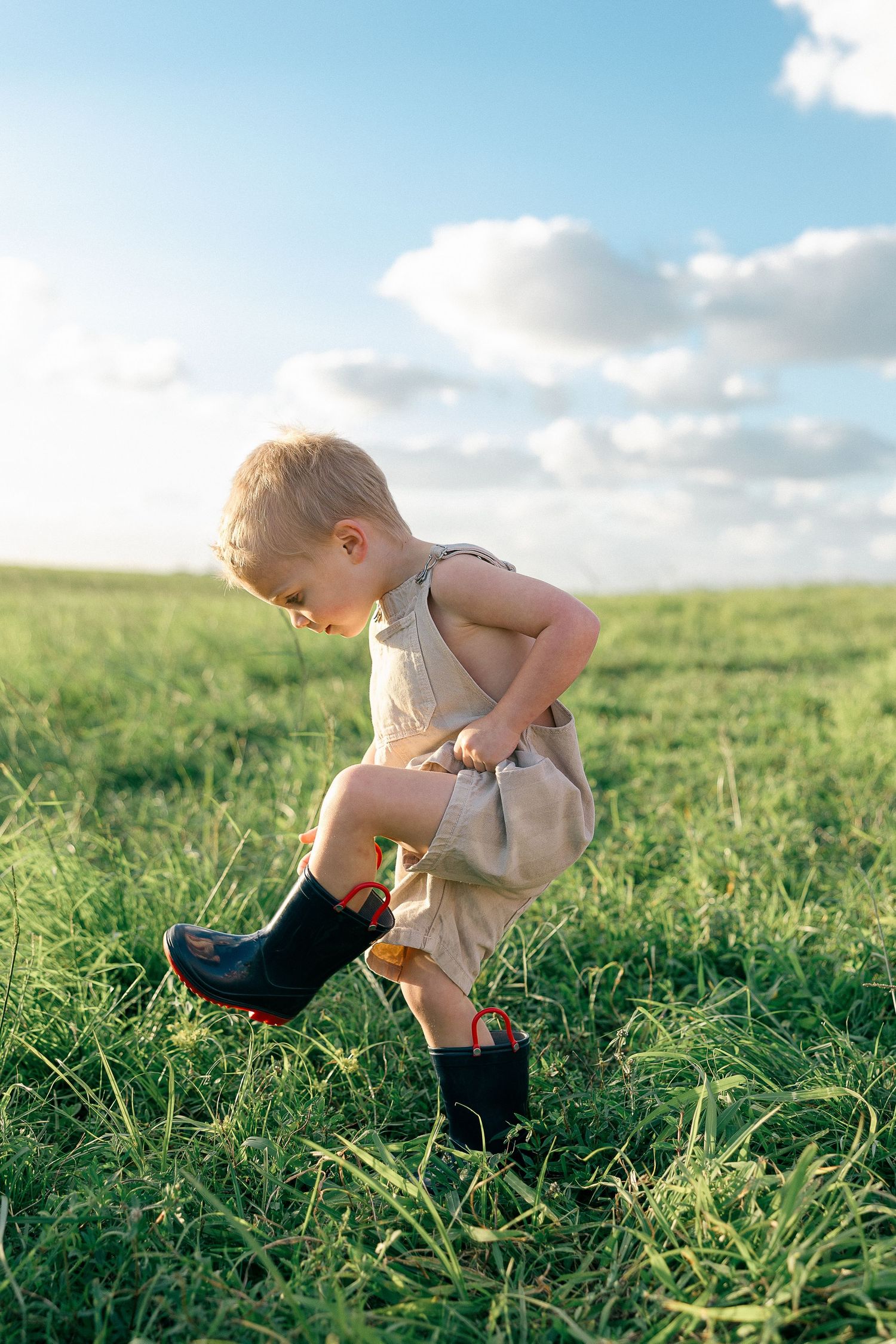 Toddler in linen romper and rubber boots playing in green grass field under blue sky.