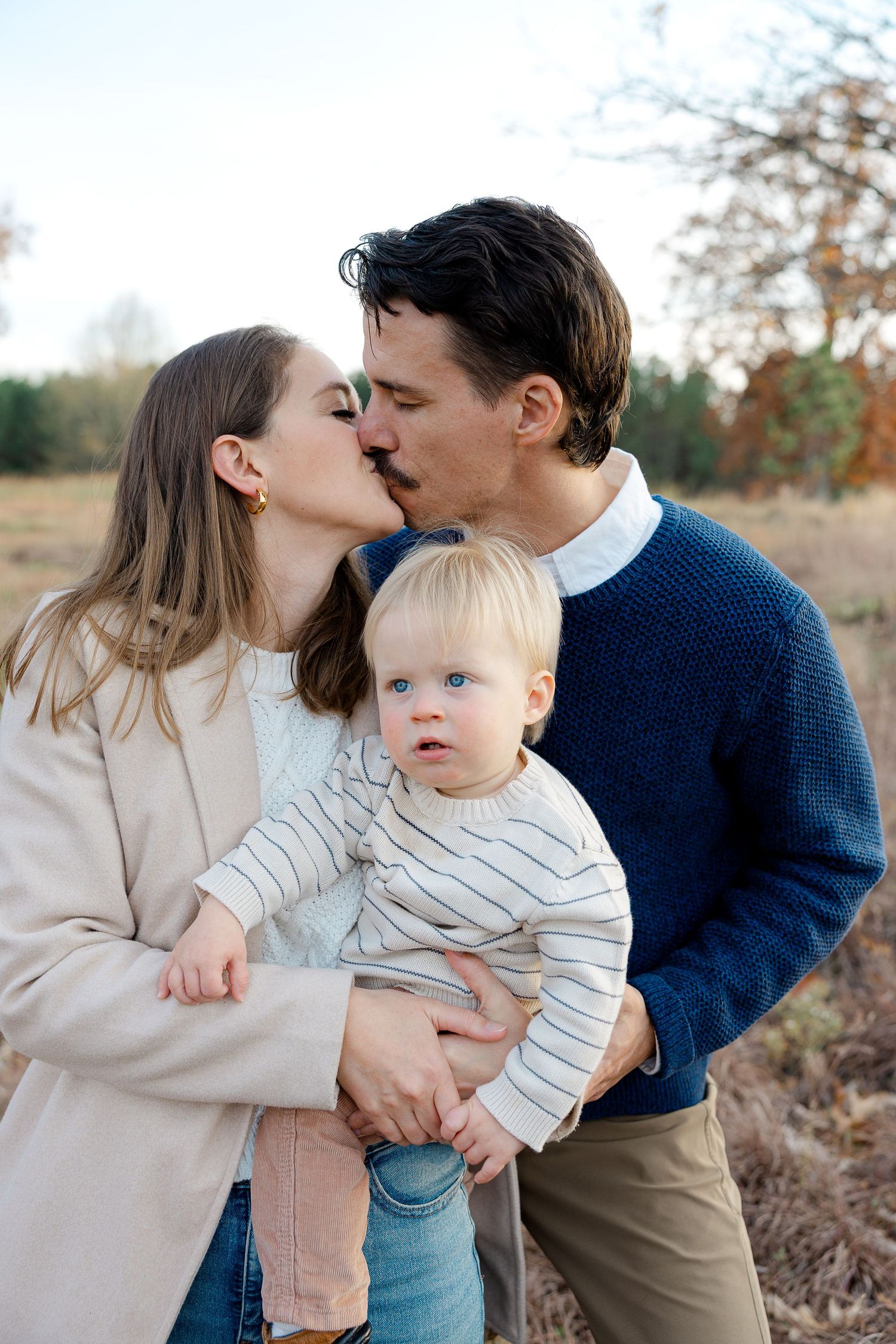A couple shares a kiss while holding their blue-eyed baby outdoors in a field.  | Karina Thomson Photography Raleigh NC