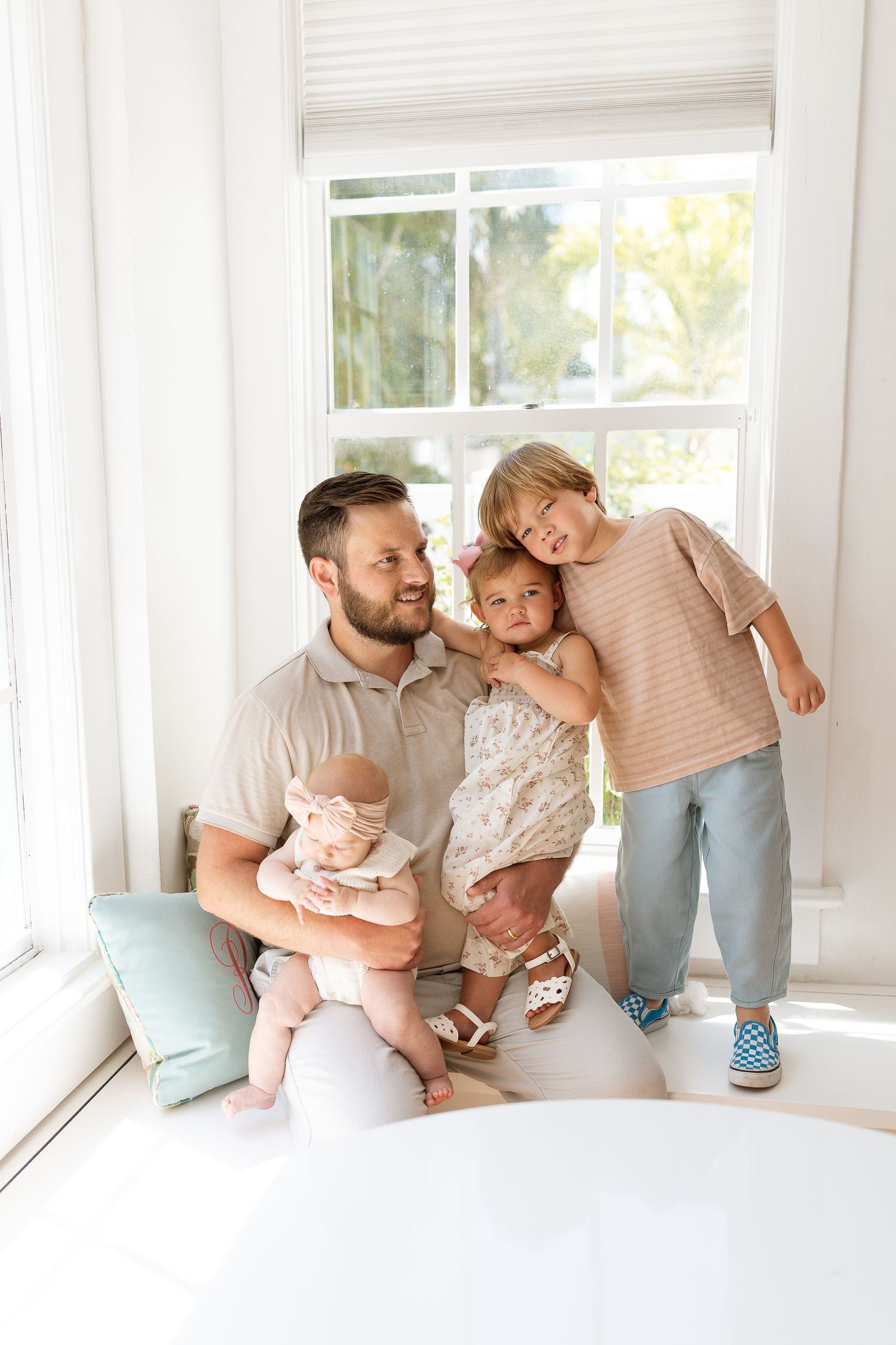 A father sits by a bright window holding a baby while two young children smile and lean against him.