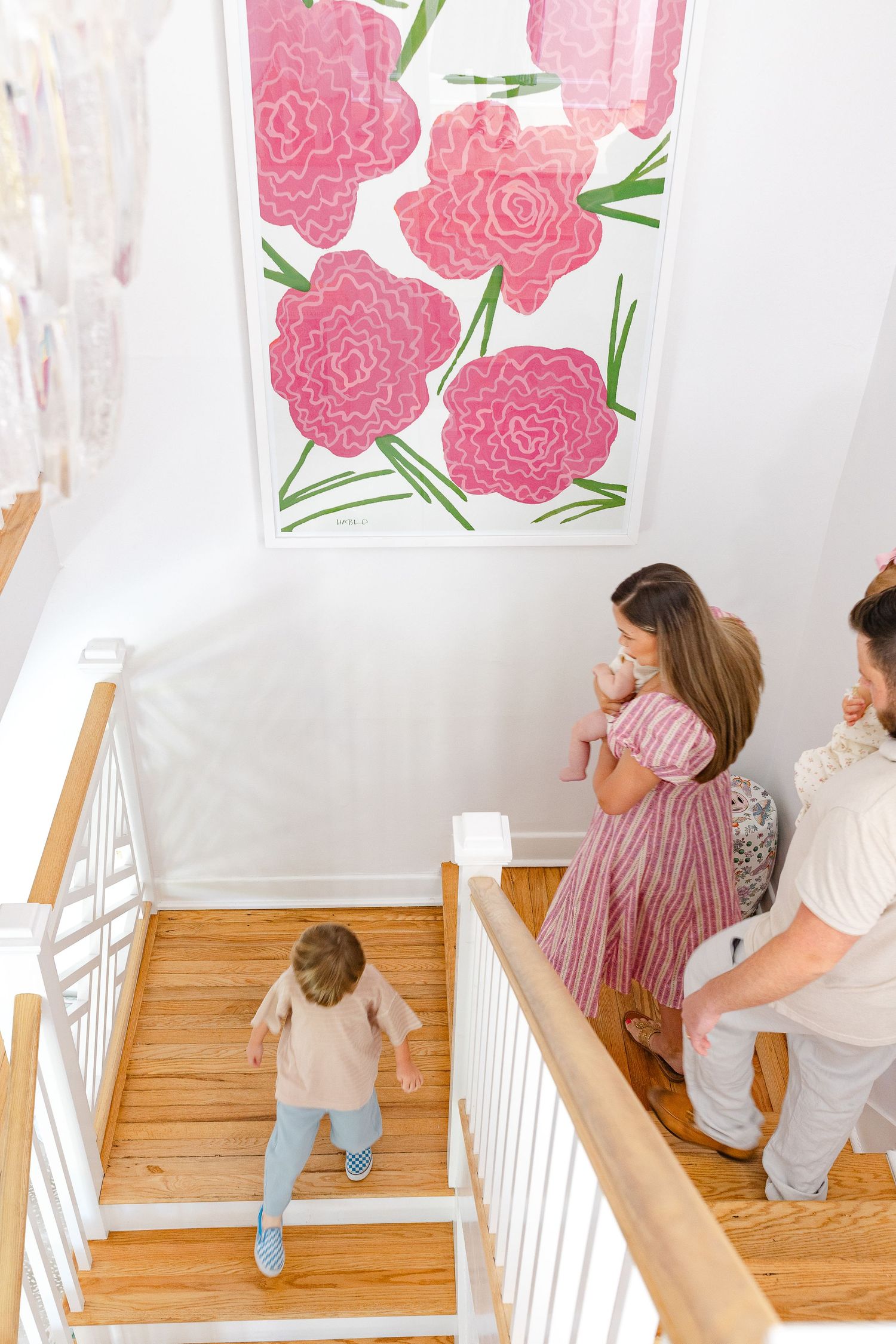 Family on wooden staircase with large pink floral wall art, toddler walking up steps in bright white home interior.