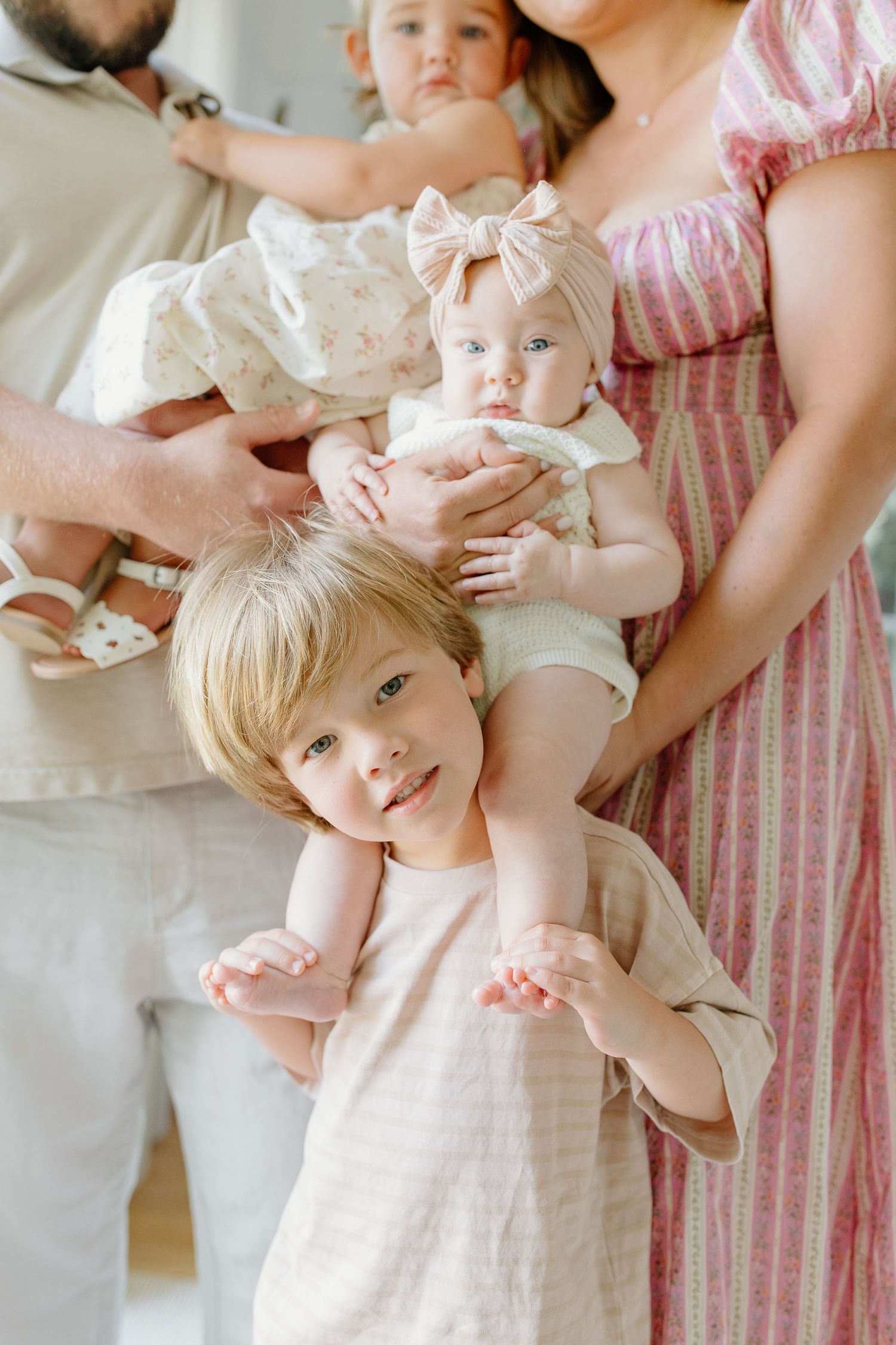 Young blonde toddler poses in front of parents holding two babies during a bright family portrait session.