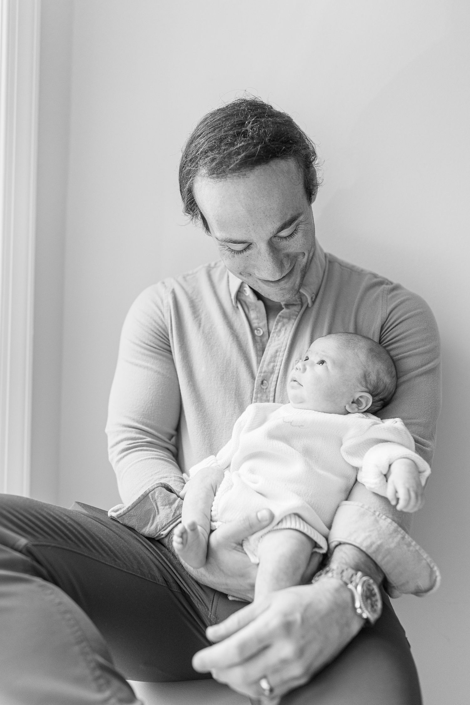 Black and white photo of a smiling father cradling his newborn baby while seated near a window.  | Karina Thomson Photography Raleigh NC