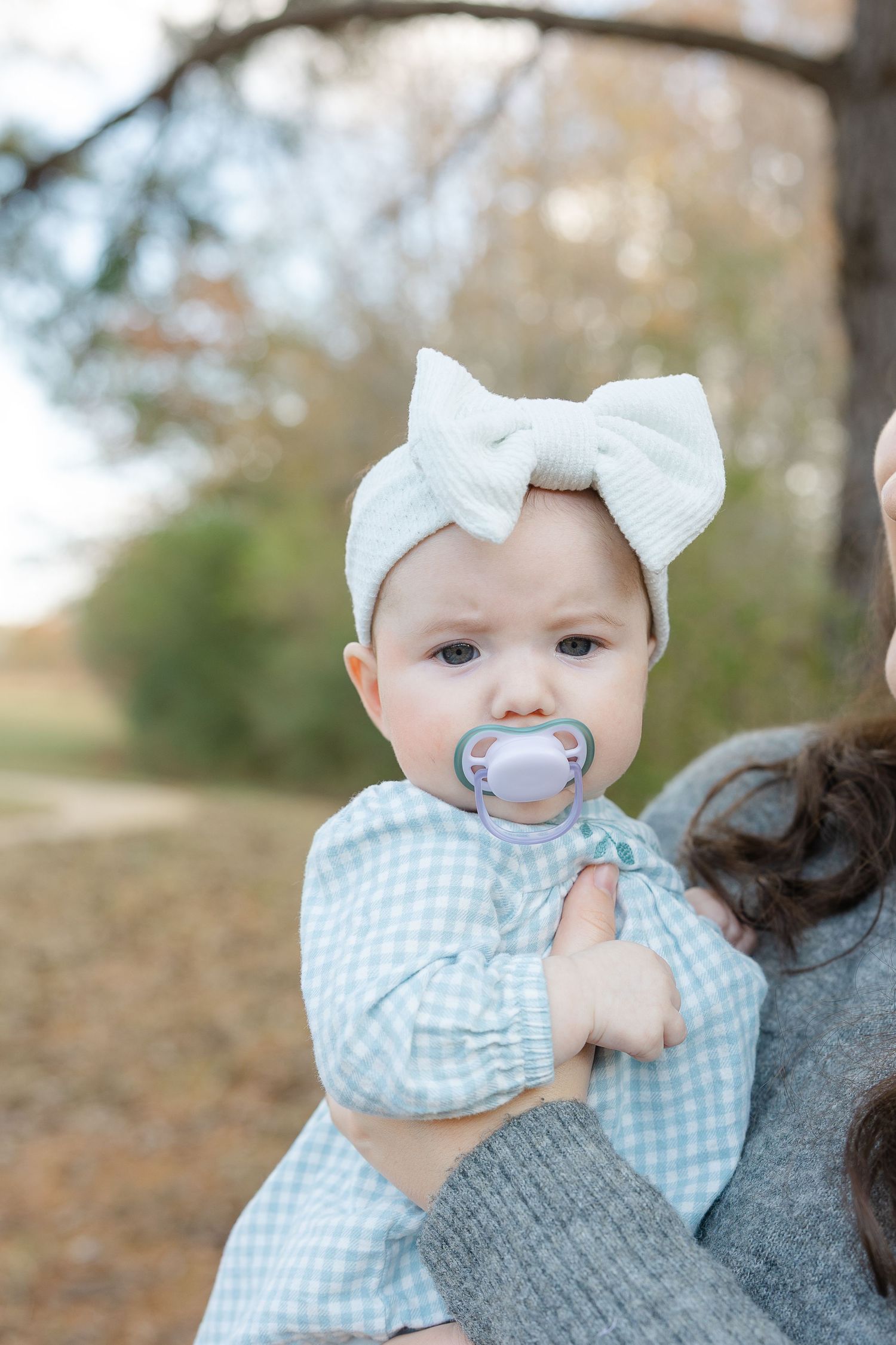 Adorable baby girl with white bow headband and pacifier being held outdoors in autumn park.  | Karina Thomson Photography Raleigh NC