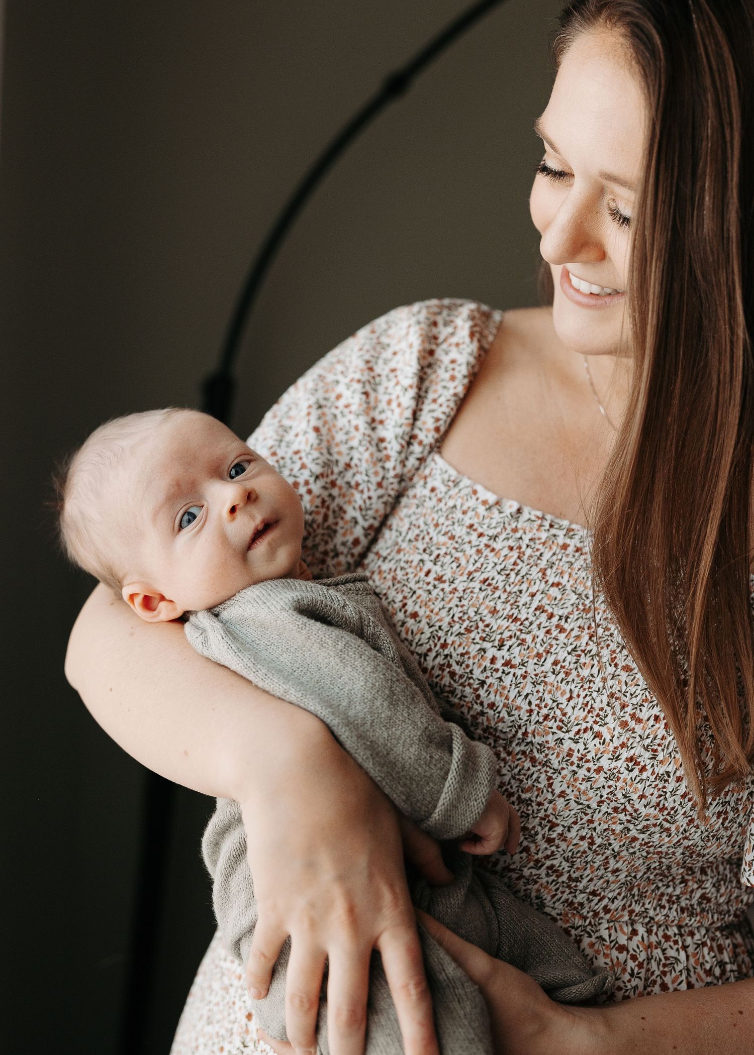 Smiling mother in a floral dress gazing lovingly at her alert newborn baby in her arms.  | Karina Thomson Photography Raleigh NC