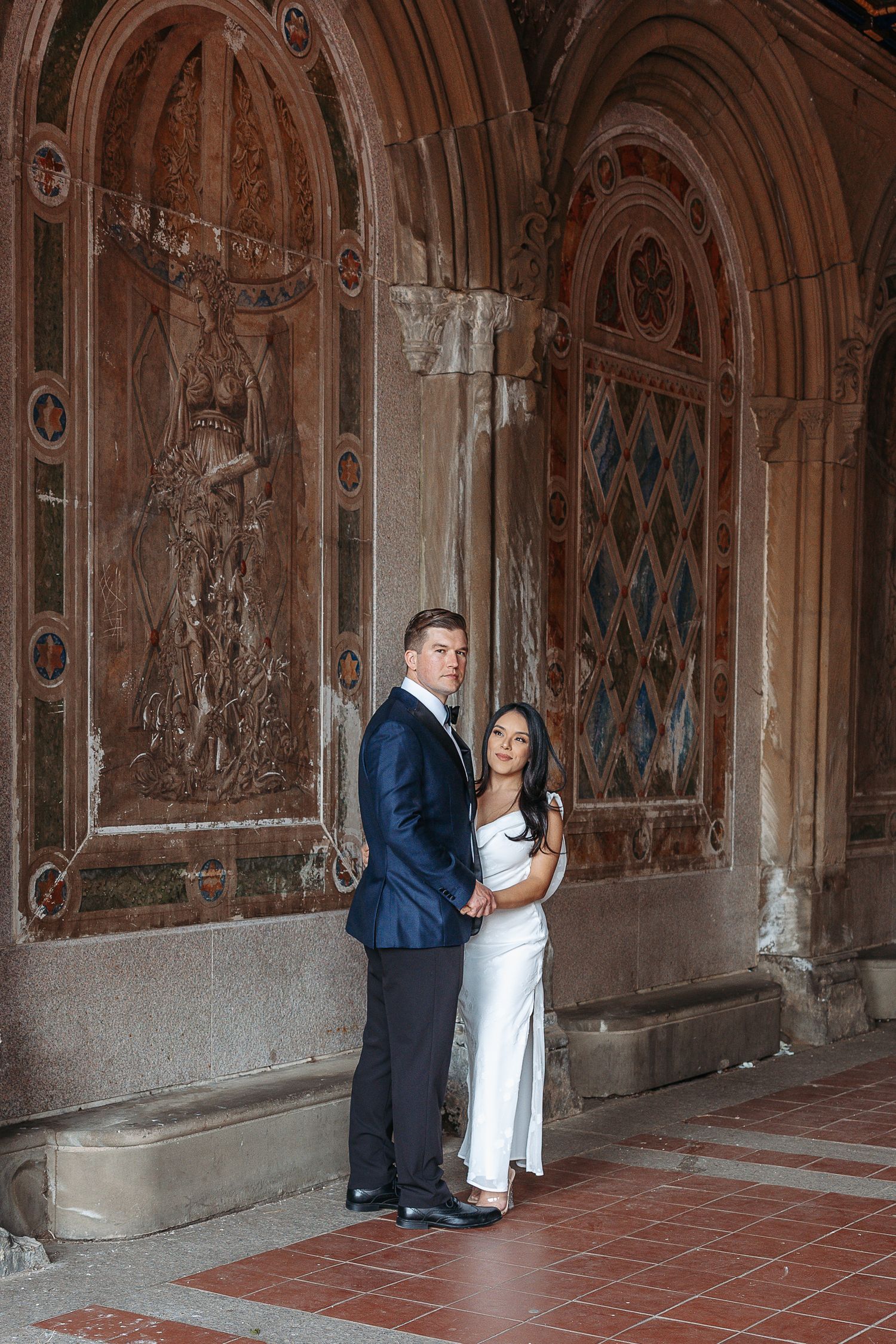 Elegant couple in formal wear embracing before ornate stone arch in Central Park Bethesda Terrace.  | Karina Thomson Photography Raleigh NC