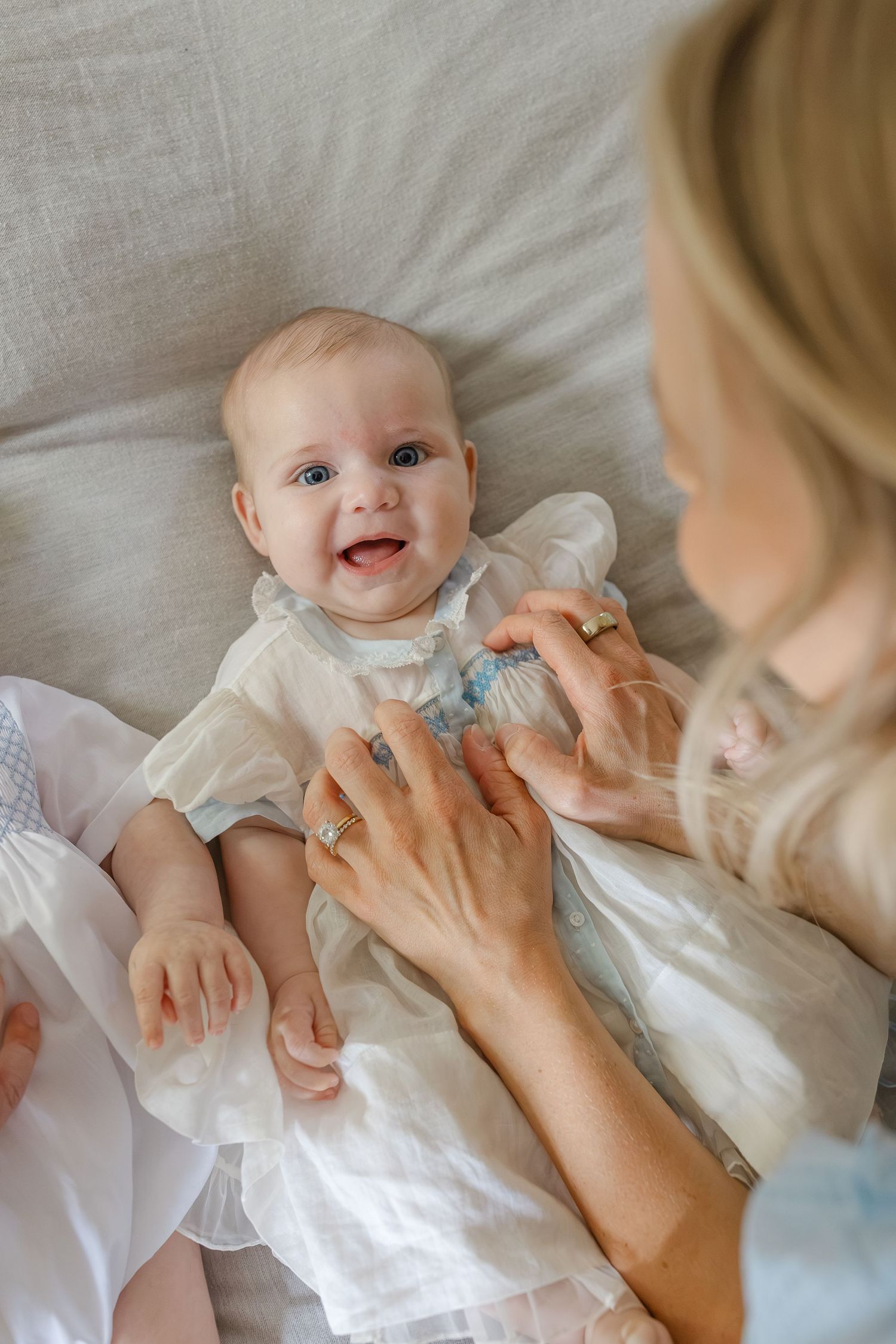 Happy baby in white dress smiling at blonde mother during a tender moment on a light gray sofa.