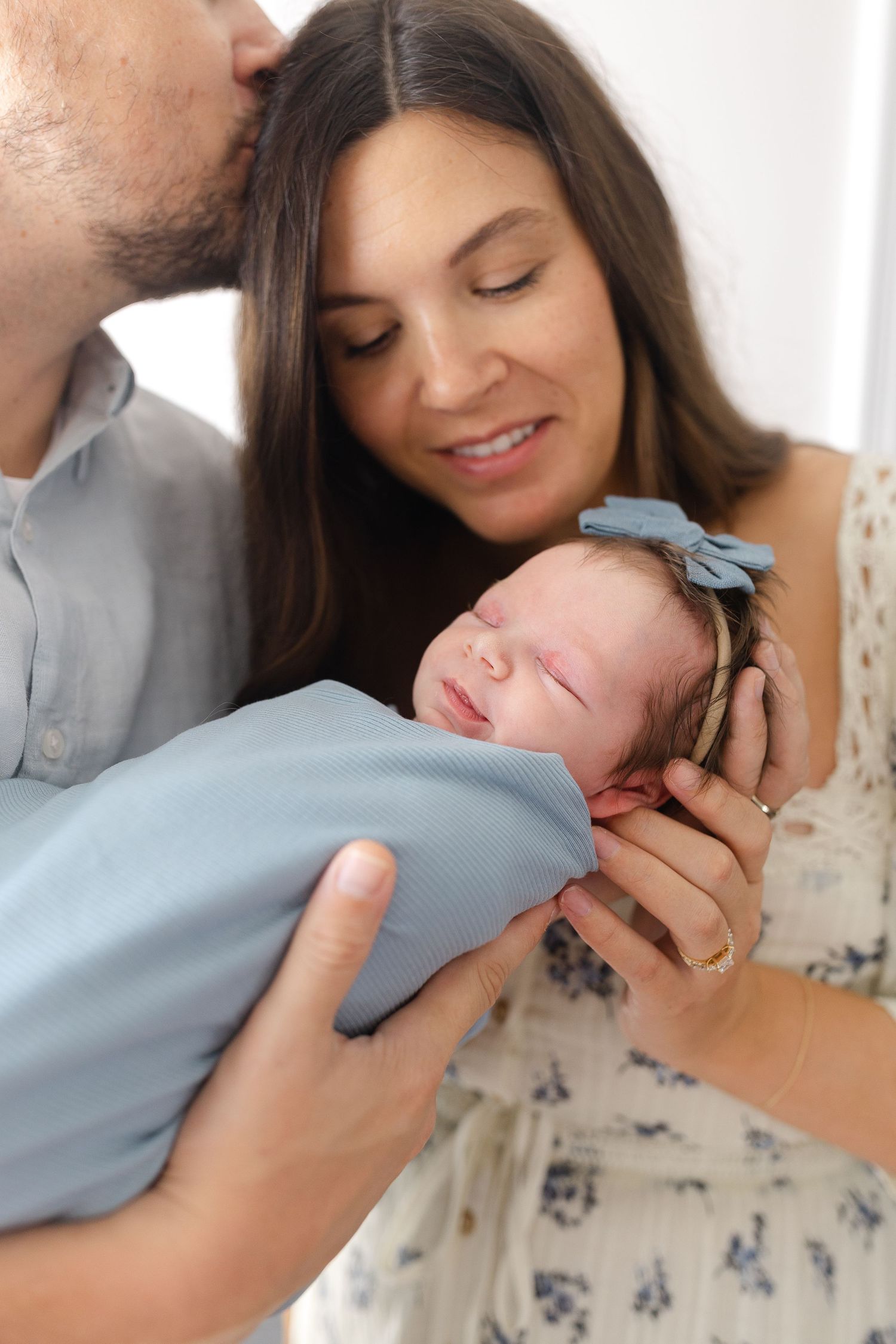 Parents tenderly hold and kiss their swaddled newborn baby wrapped in a blue blanket.  | Karina Thomson Photography Raleigh NC