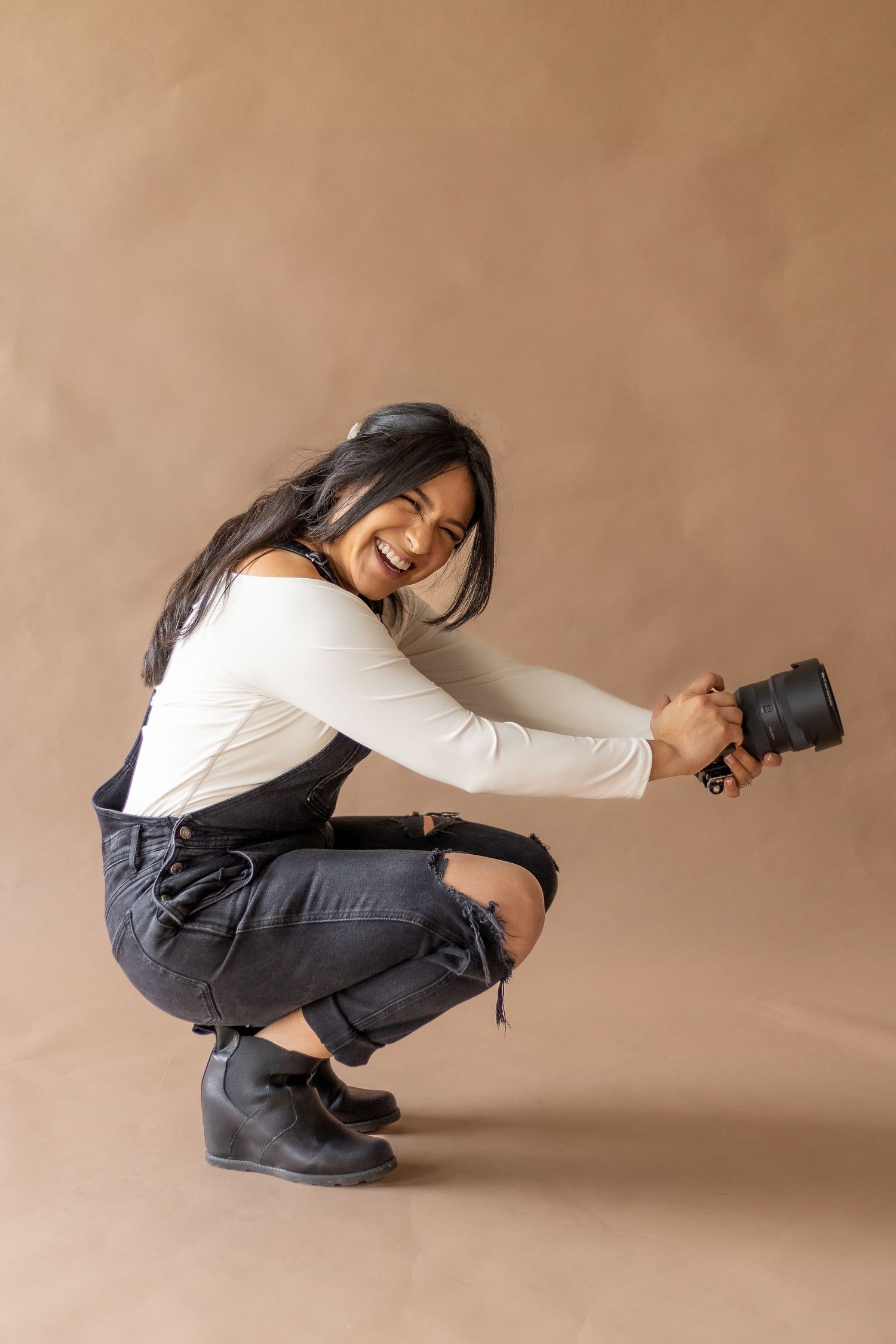 Laughing female photographer crouches holding camera against a warm brown background.  | Karina Thomson Photography Raleigh NC