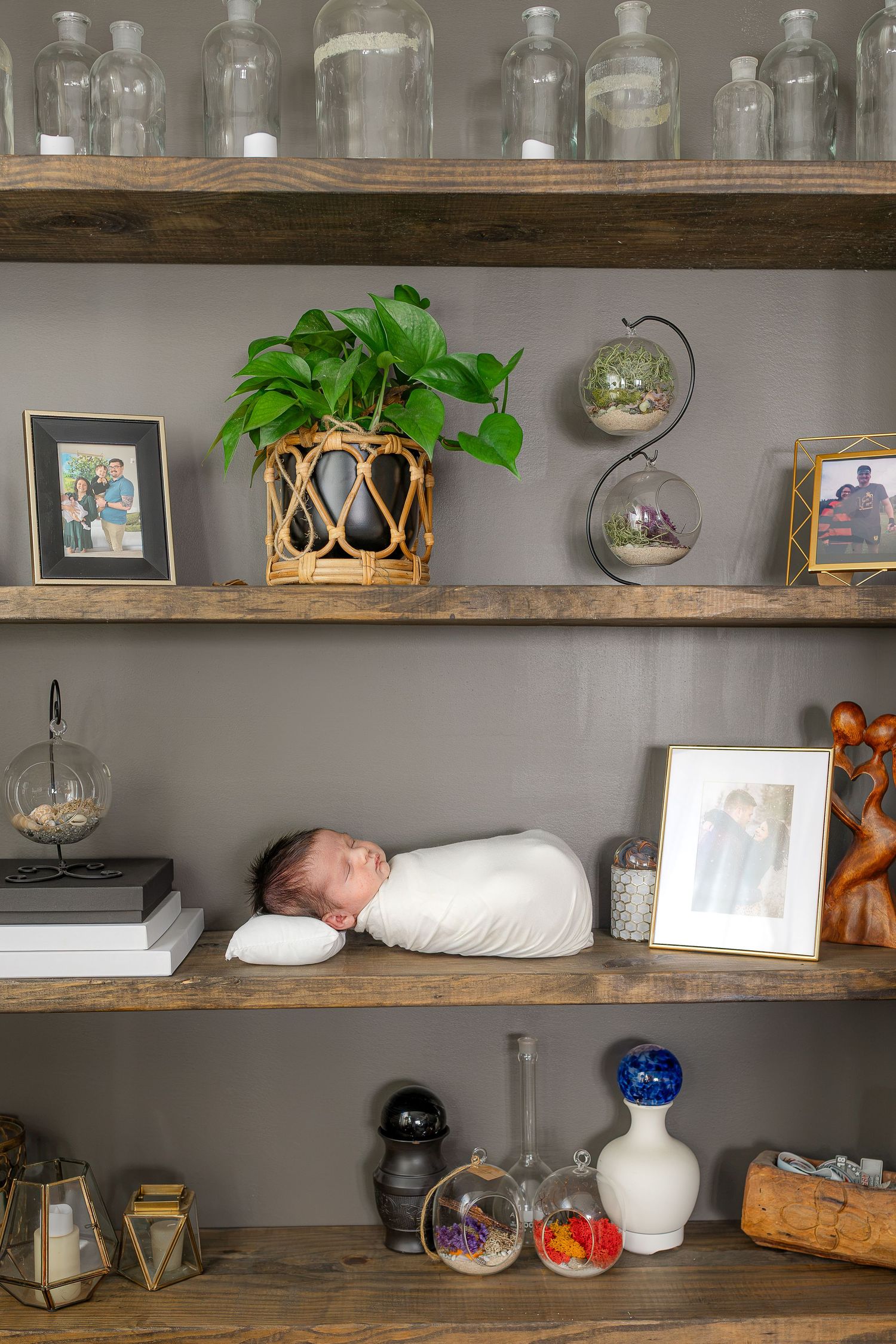 Newborn baby swaddled in white, sleeping peacefully on a wooden shelf surrounded by plants, frames, and decorative items.
