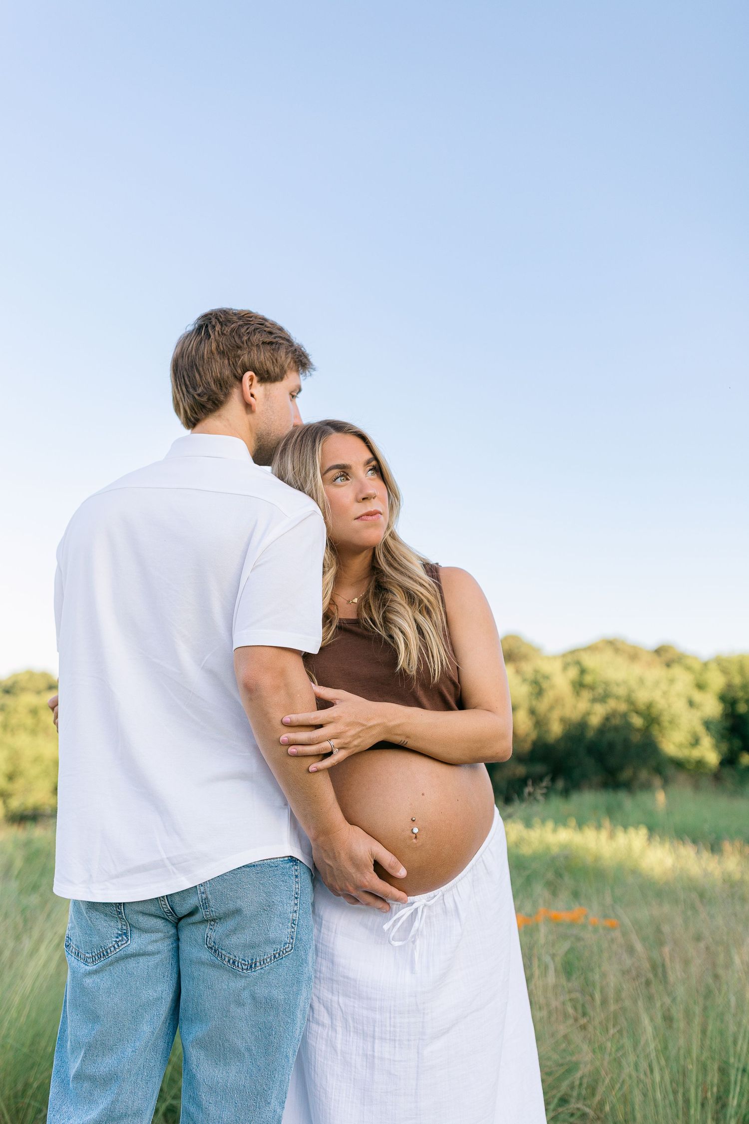 Pregnant couple embracing in a sunny green field, woman in brown top and white skirt cradling her baby bump.