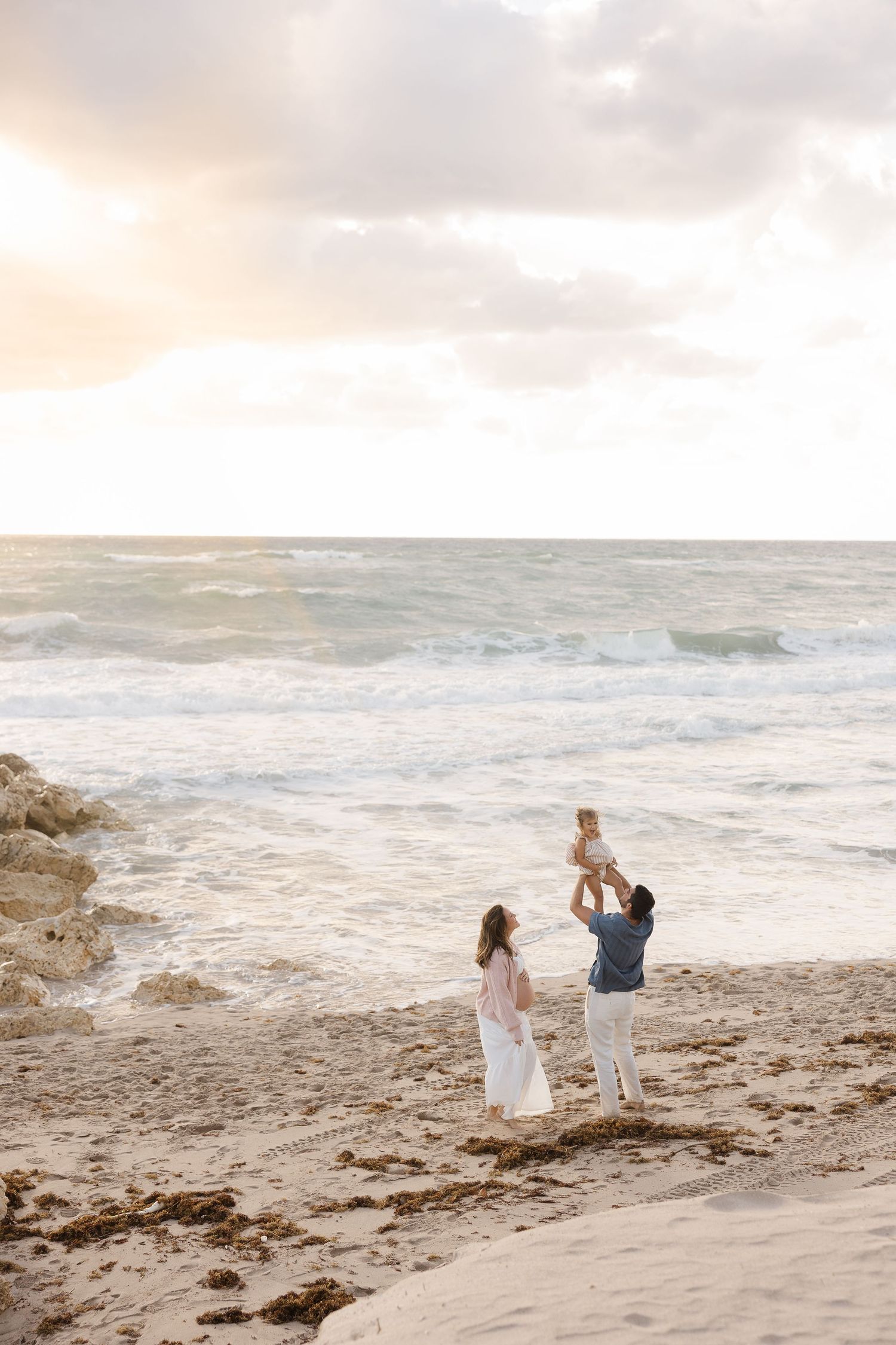 Parents stand on sandy beach at sunset as father lifts baby high in the air near ocean waves.  | Karina Thomson Photography Raleigh NC