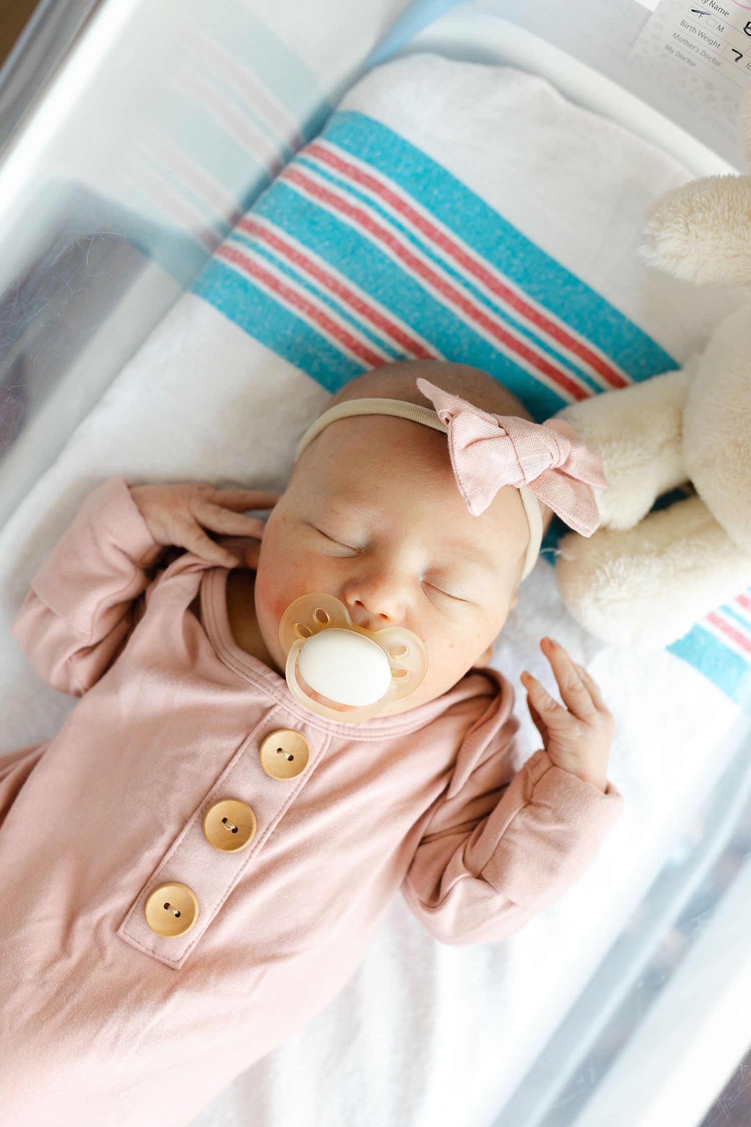 Newborn baby girl sleeping peacefully in hospital bassinet wearing pink outfit with bow headband and white pacifier.