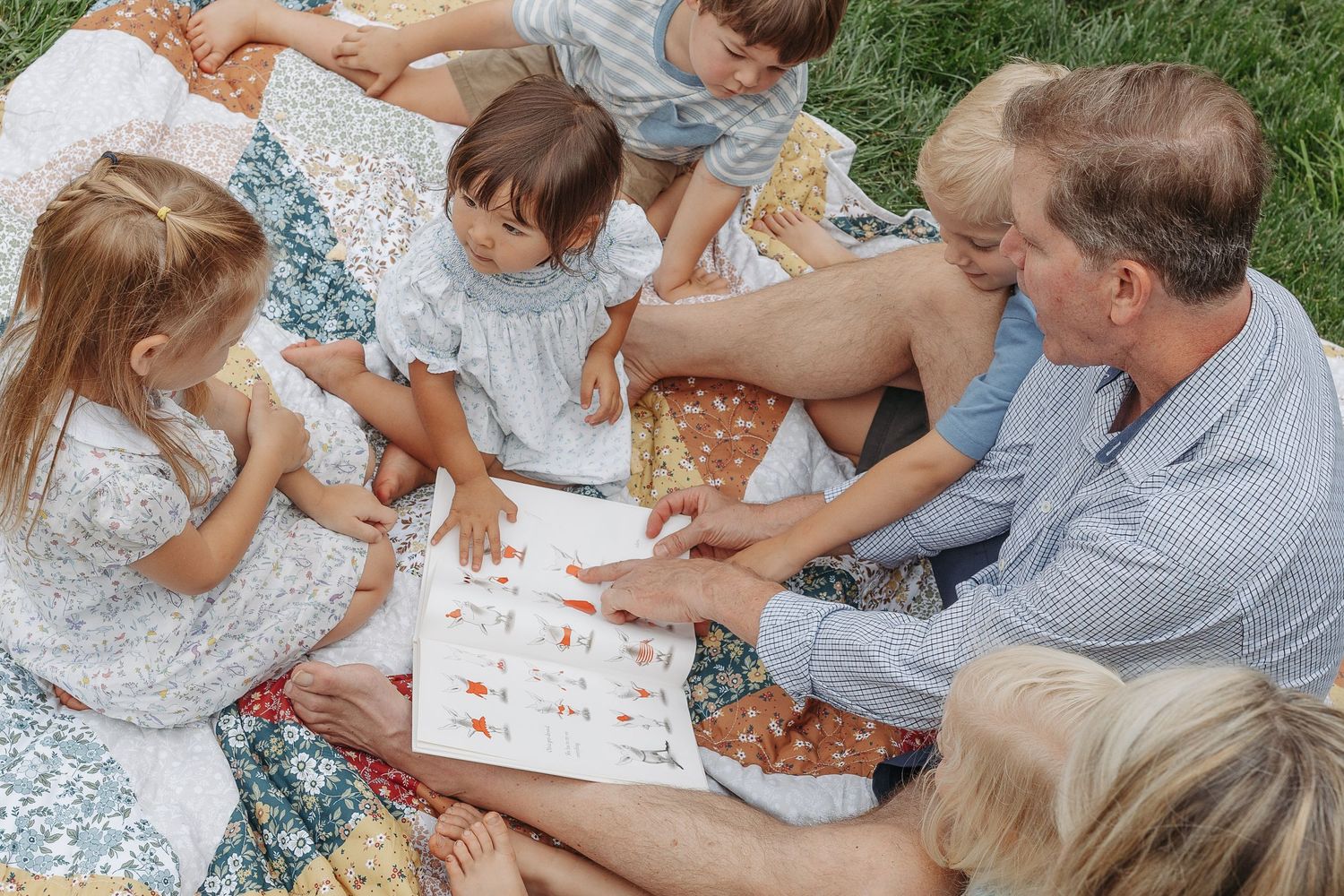 A man reads a picture book with a group of young children sitting on a colorful quilt outdoors on green grass.