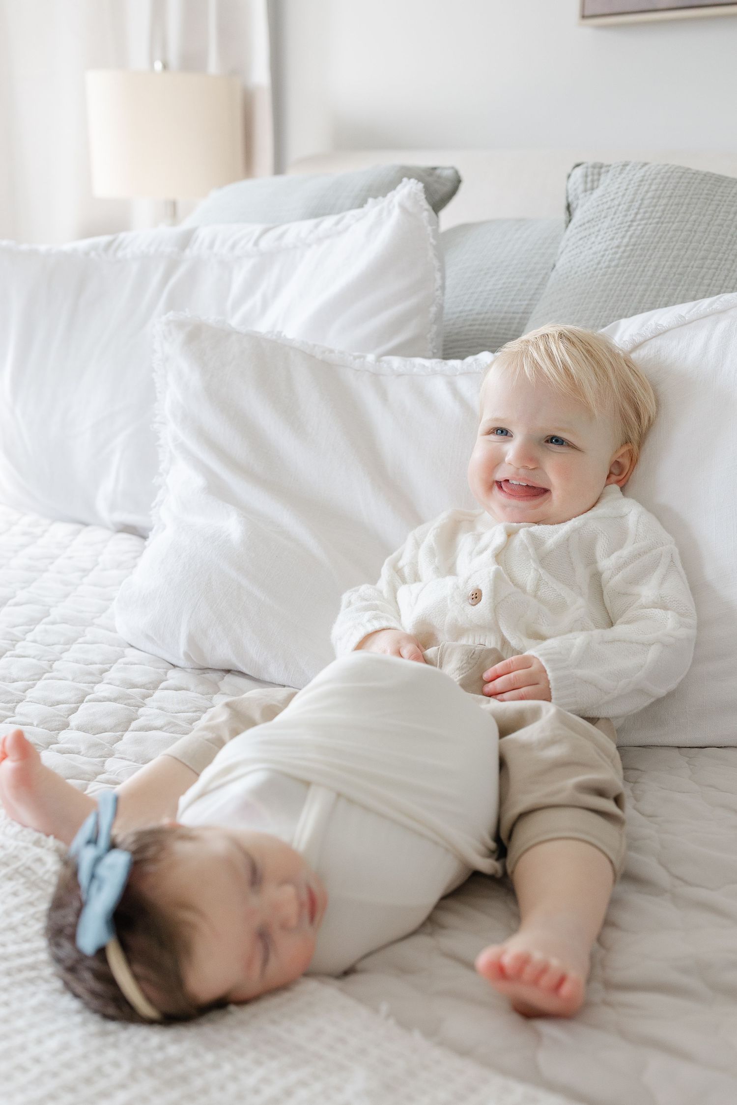 Smiling toddler sits beside sleeping newborn with blue bow on a white bed.  | Karina Thomson Photography Raleigh NC