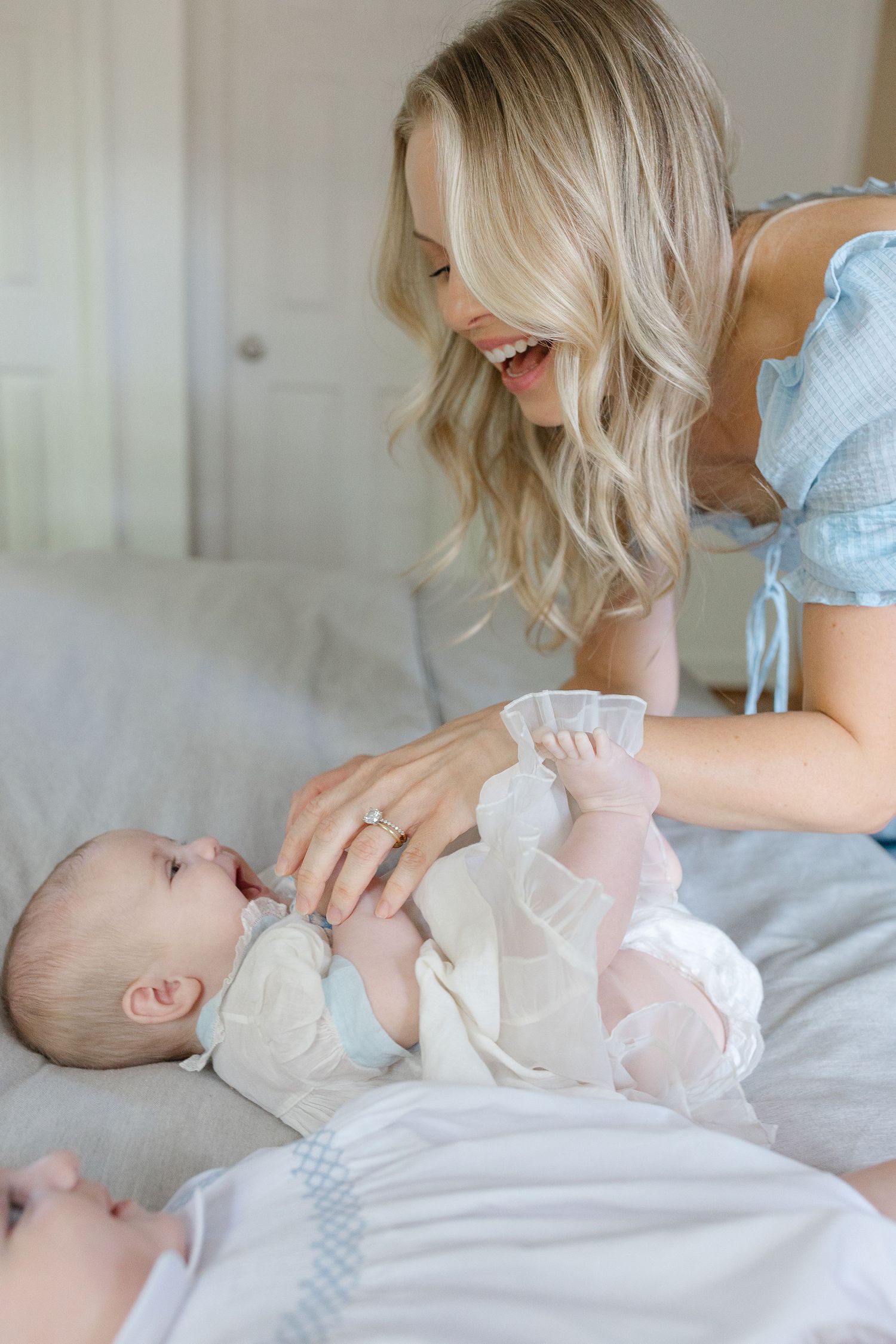 Smiling blonde mother playing with her newborn baby lying on a bed, wearing a light blue dress.