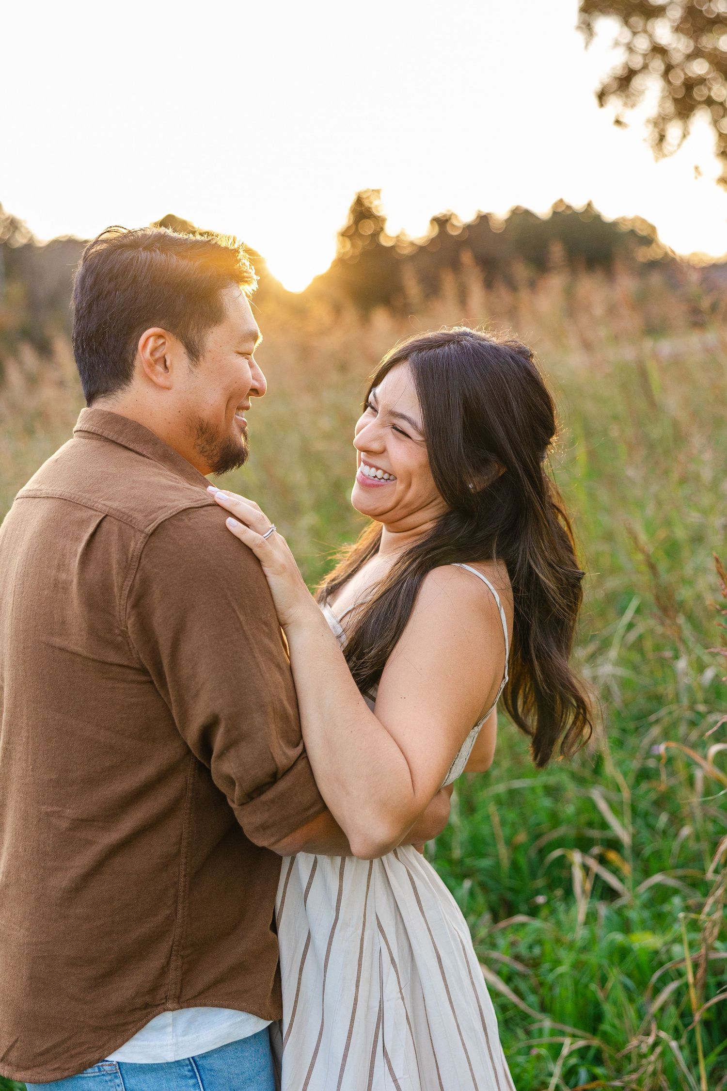 Couple embracing and laughing in a golden sunset field, woman in striped dress holding man in brown shirt.
