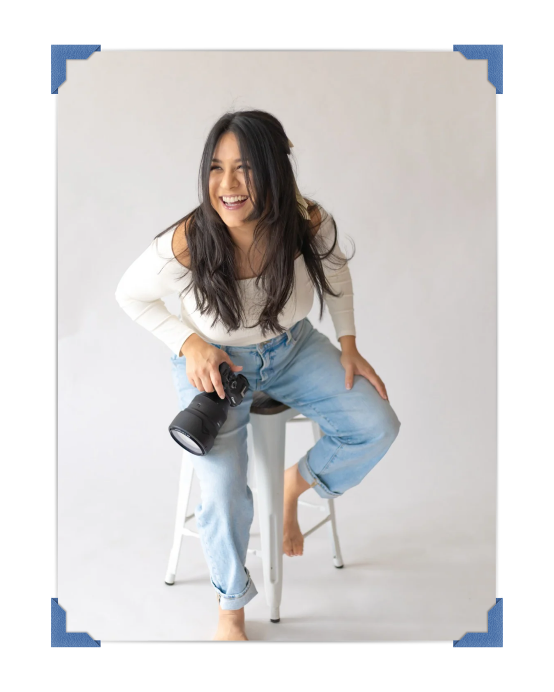Smiling female photographer sits on a stool holding a camera in a bright white studio.  | Karina Thomson Photography Raleigh NC