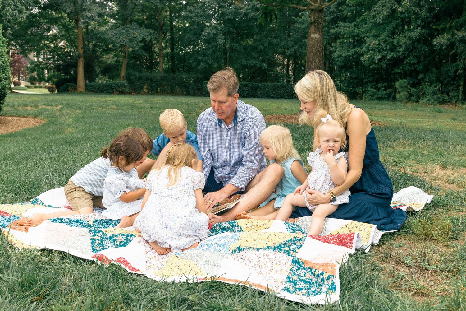 Family with five young children sitting on a colorful patchwork quilt outdoors, reading together in a lush green yard.