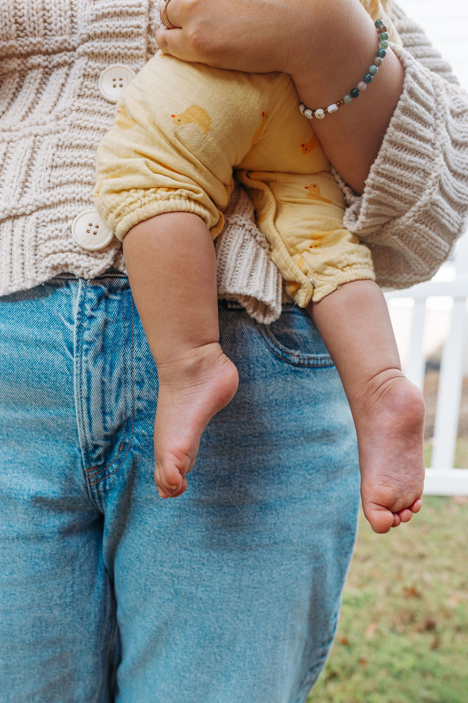 Close-up of baby feet dangling as mother in jeans and knit cardigan holds infant outdoors.  | Karina Thomson Photography Raleigh NC