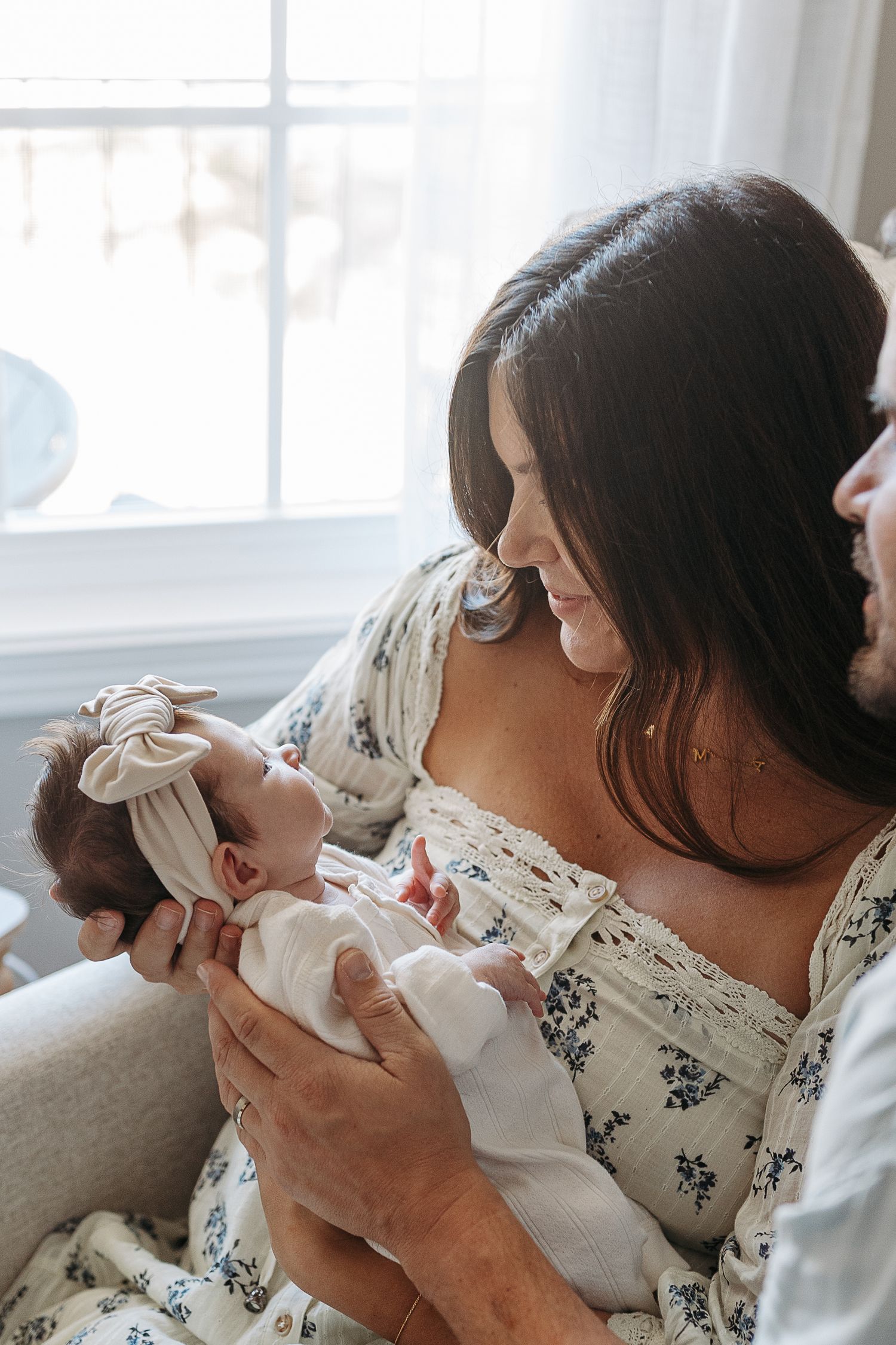 Mother holds newborn baby while father looks on lovingly near a sunlit window.  | Karina Thomson Photography Raleigh NC