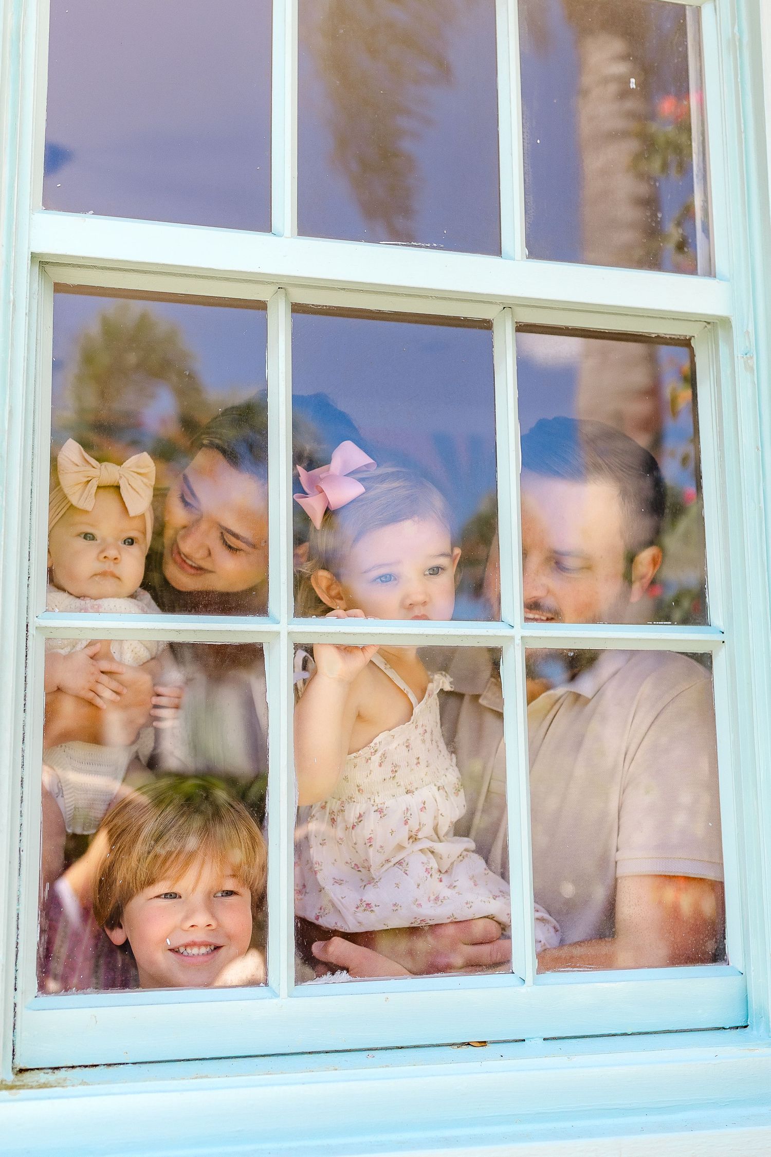 Family of four smiling through a white-framed window, with two young girls wearing pink bows and a baby.