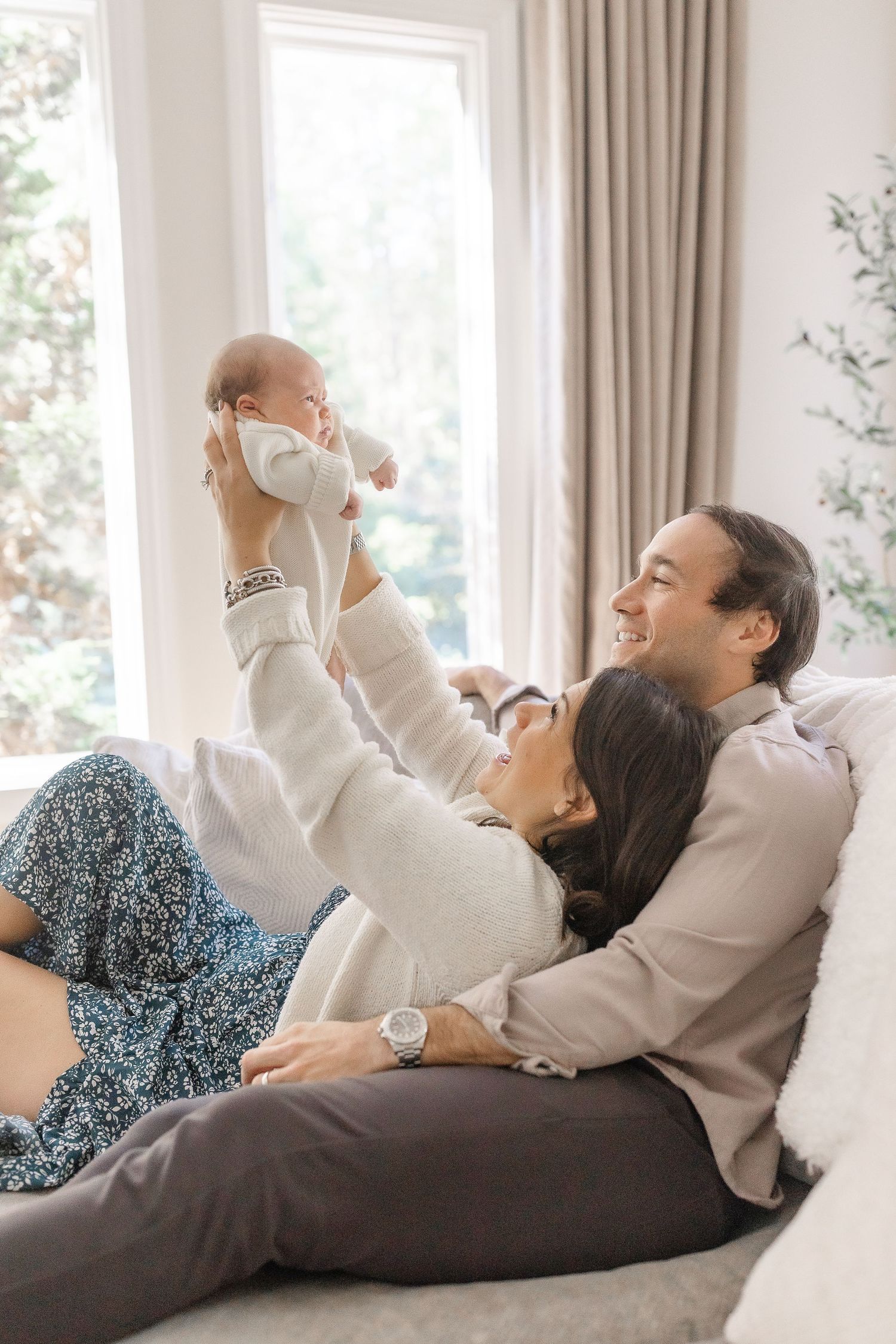 Happy couple on a sofa as mother lifts their smiling newborn baby joyfully in the air.  | Karina Thomson Photography Raleigh NC