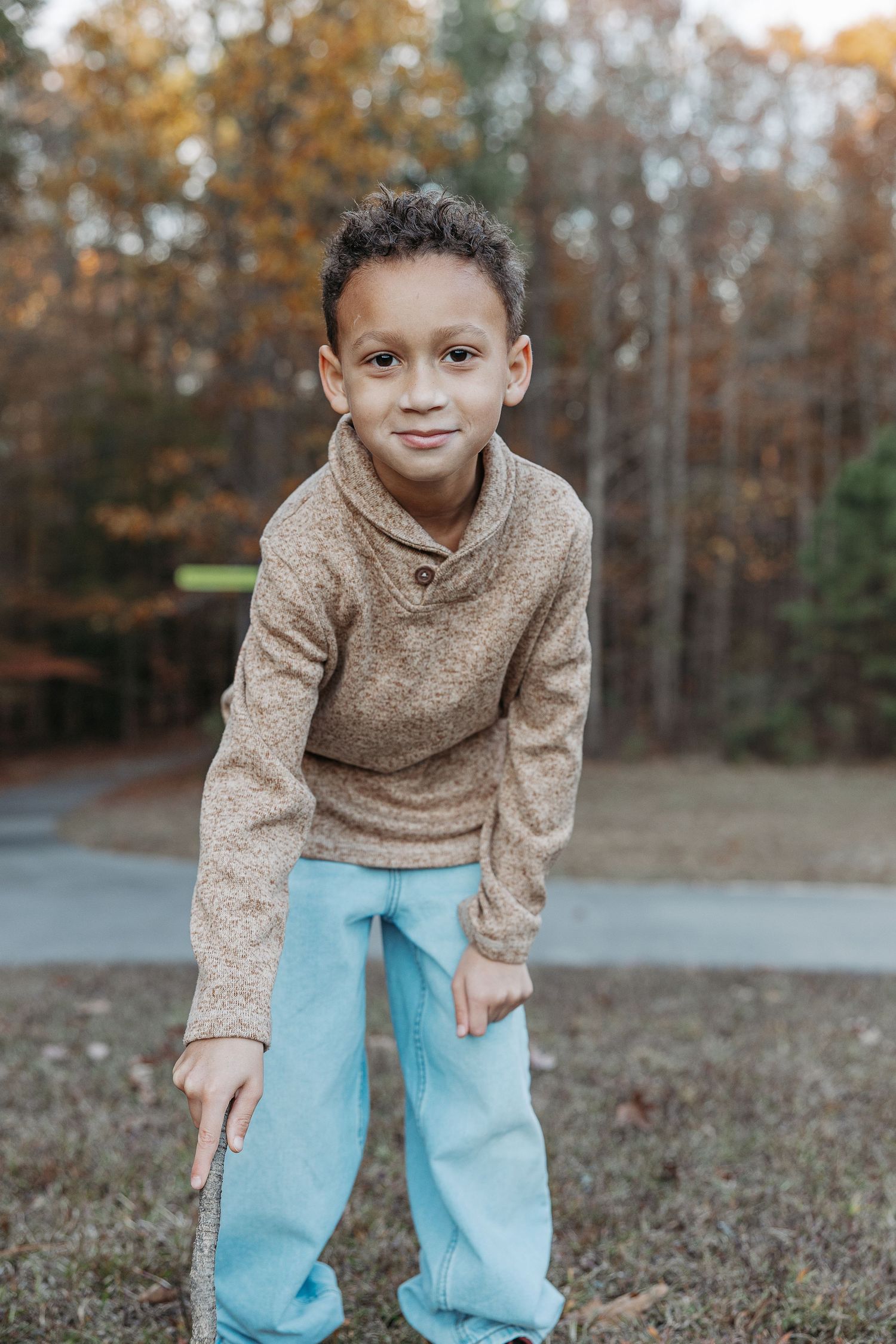 Smiling young boy in brown sweater and blue pants posing outdoors in autumn setting.  | Karina Thomson Photography Raleigh NC