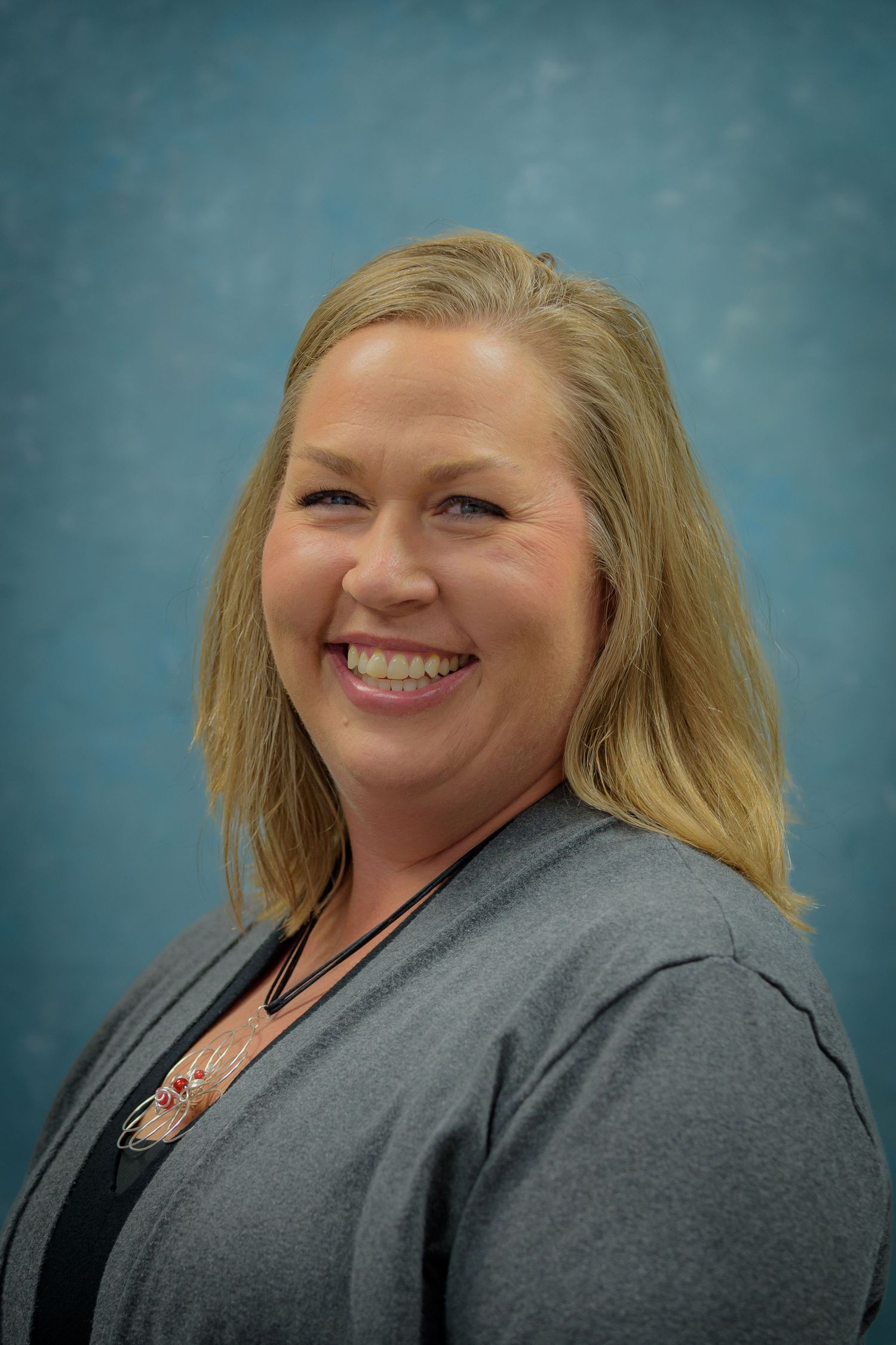 Smiling blonde woman in gray cardigan poses for professional headshot against blue background.