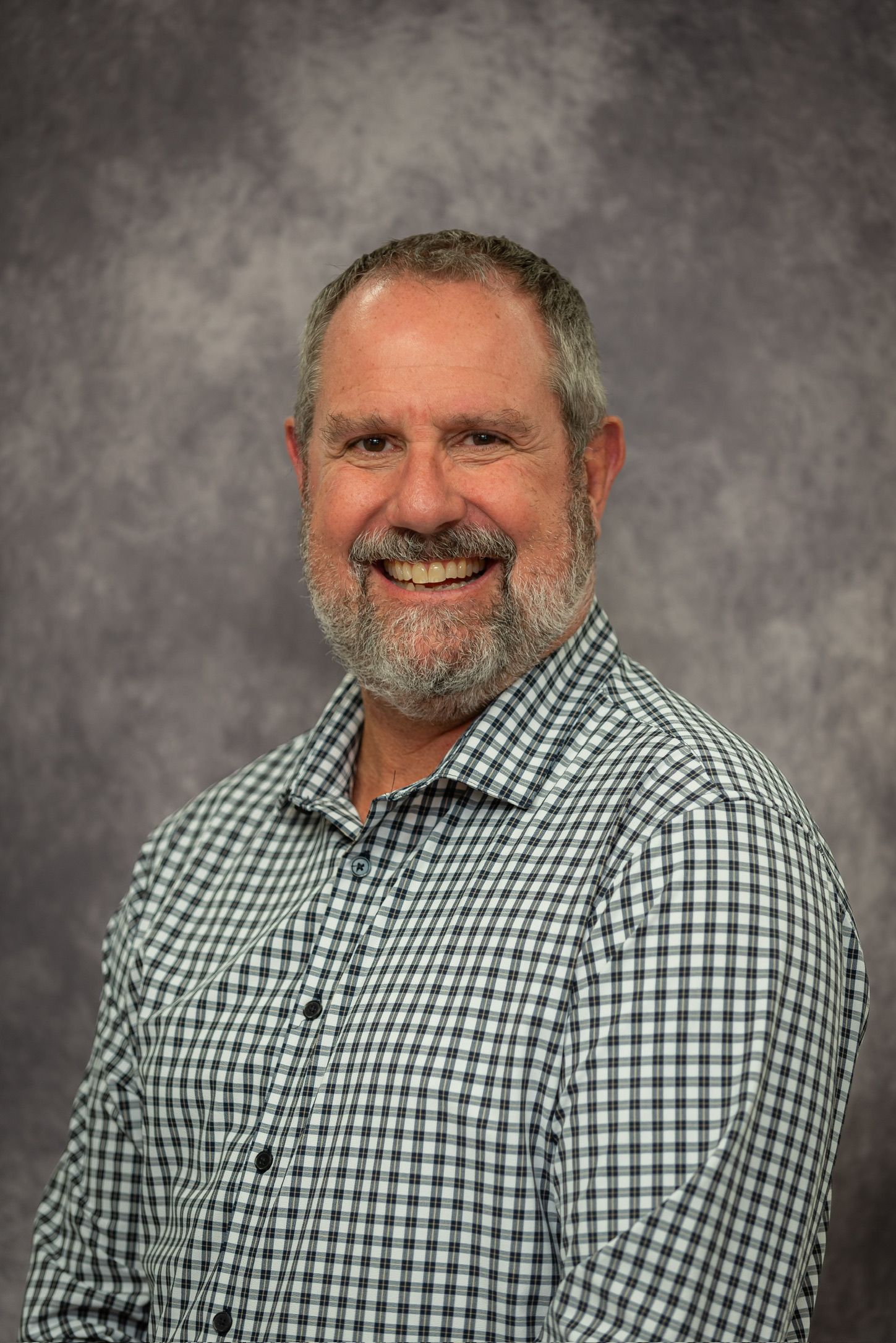 Smiling middle-aged man with gray beard wearing green plaid shirt poses for professional headshot photo.