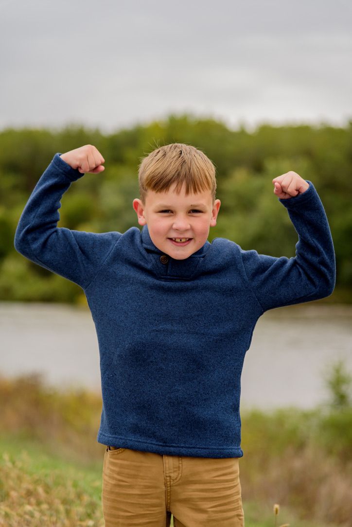 Young boy in blue sweater flexing both arms outdoors near a lake, smiling confidently.