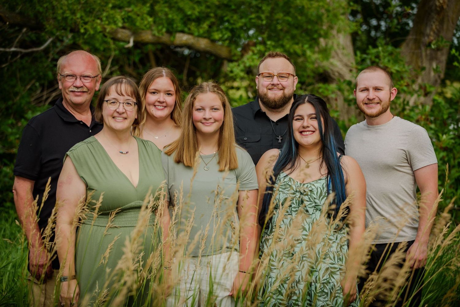Family of seven smiling together outdoors in a lush green garden setting for a portrait photo.