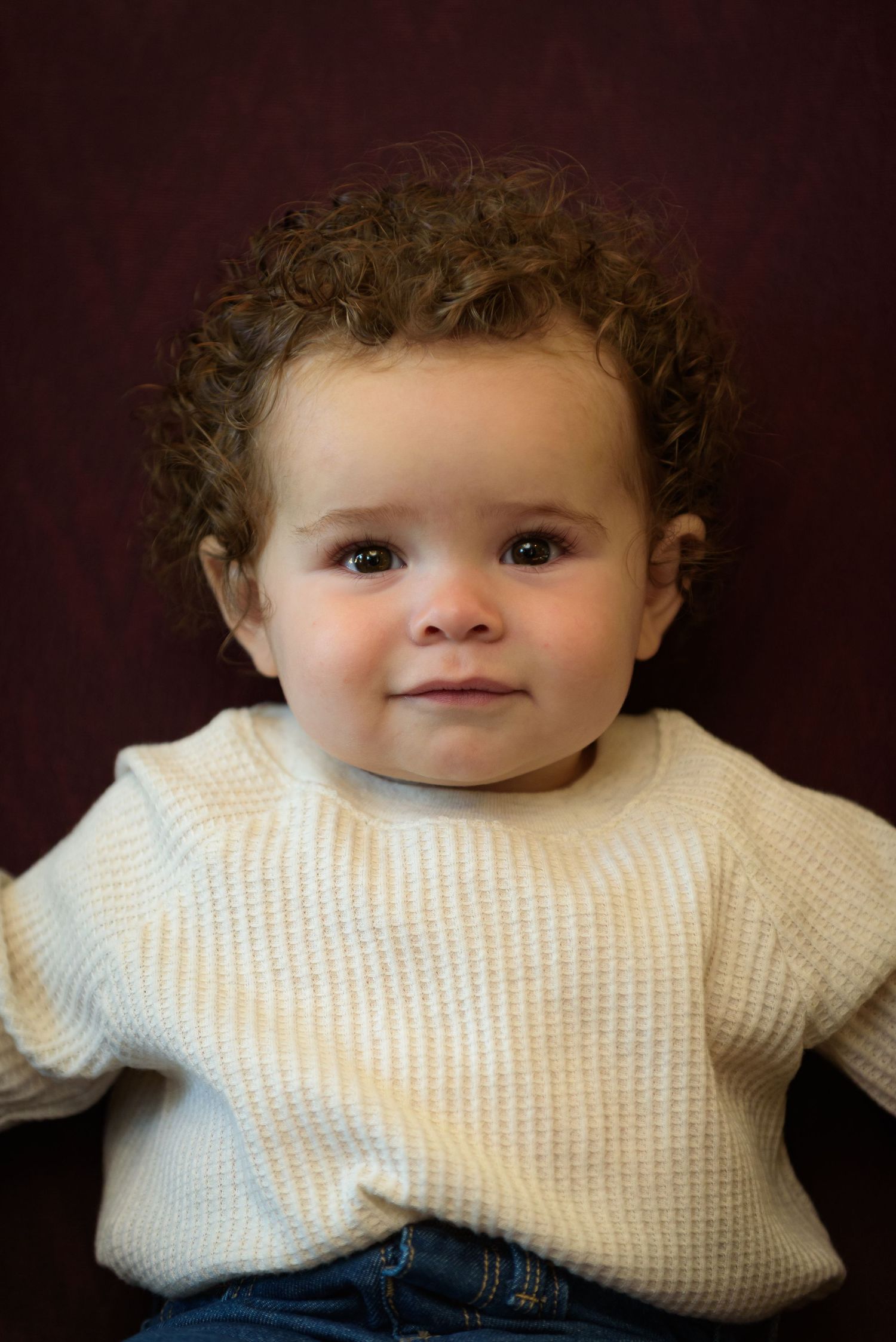 Adorable baby with curly brown hair wearing a cream knit sweater and jeans, looking at camera against dark red background.