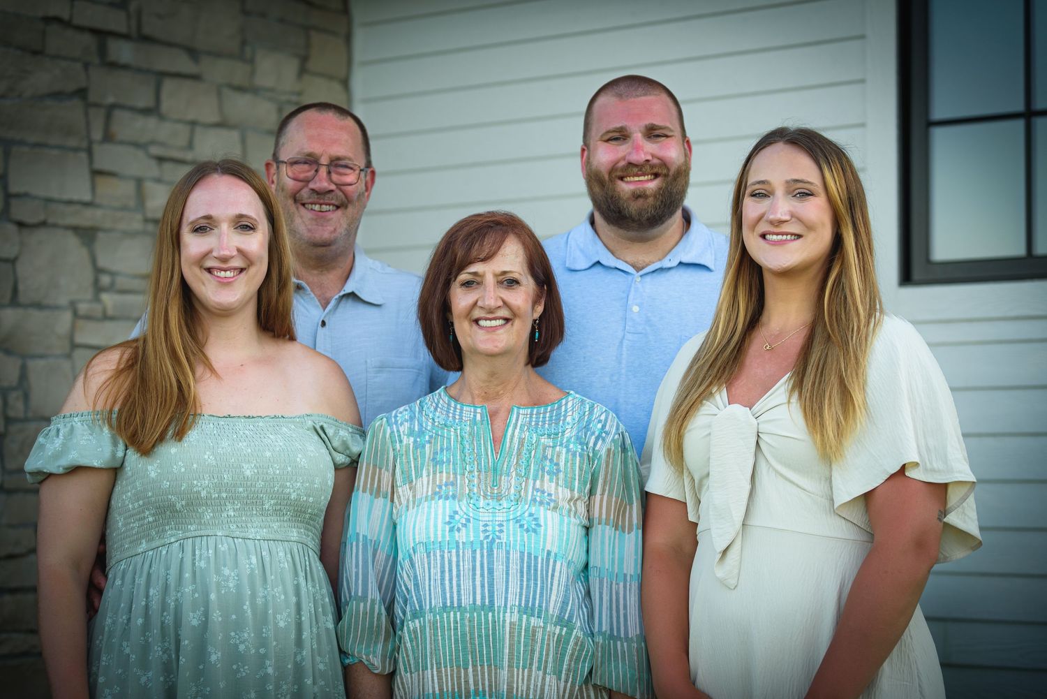 A smiling family of five poses together outdoors near a stone building for a formal portrait photo.