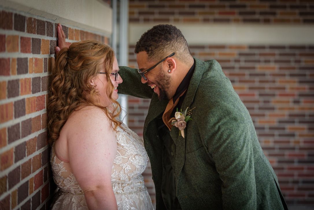 Bride and groom share an intimate moment laughing together against a brick wall on their wedding day.