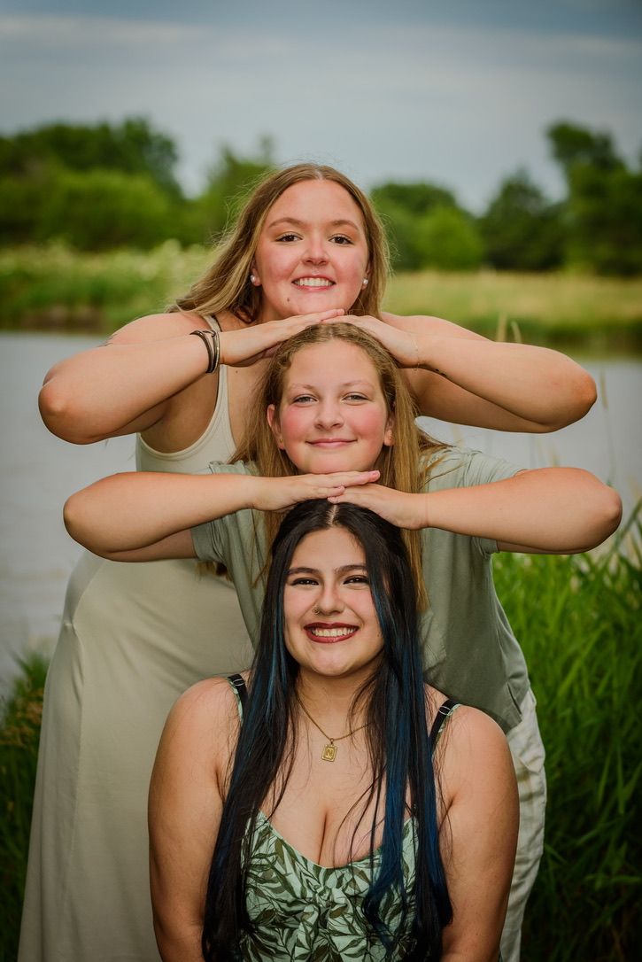 Three smiling young women pose stacked in front of a serene lake surrounded by lush green trees.