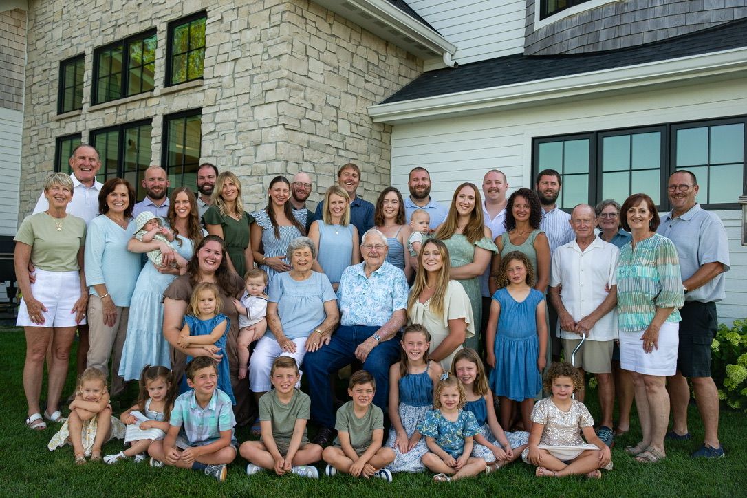 Large multigenerational family group photo outdoors in front of a stone house, dressed in coordinated blue and green outfits.