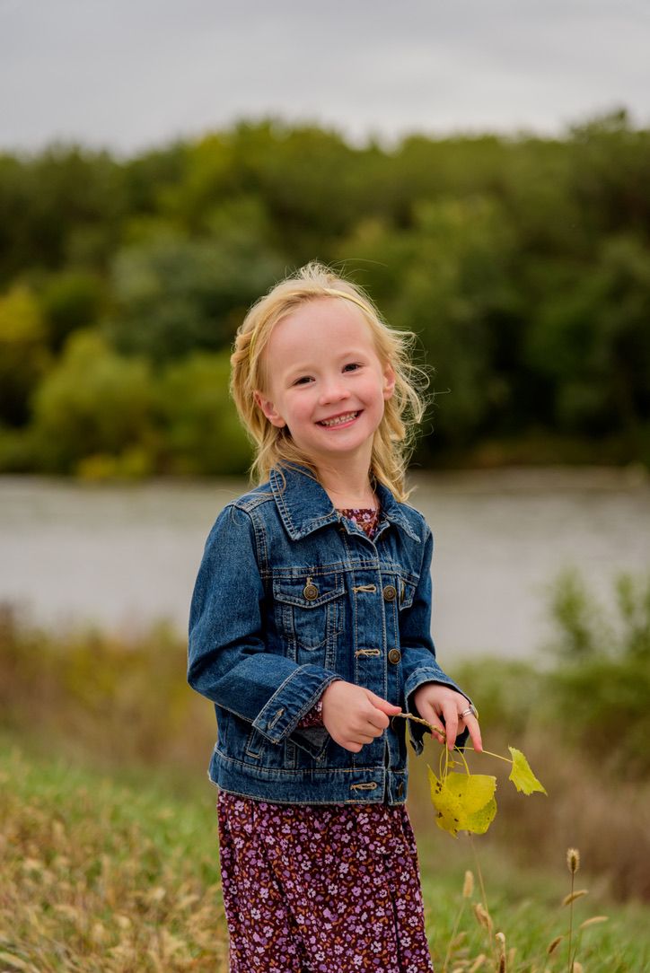 Smiling blonde girl in denim jacket holding yellow leaf outdoors near a lake in autumn.
