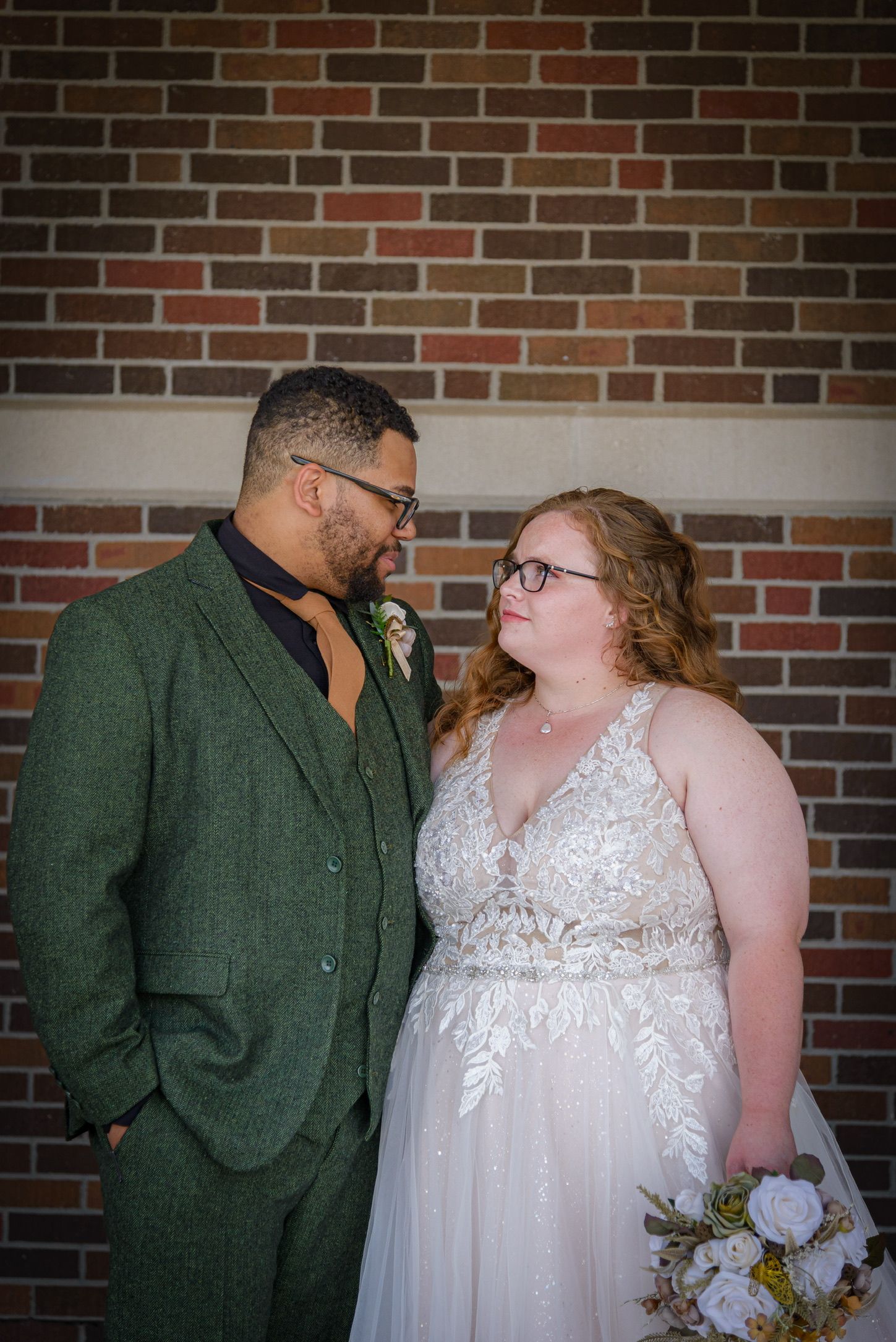 A bride in lace gown and groom in green tweed suit share a loving gaze before a brick wall.
