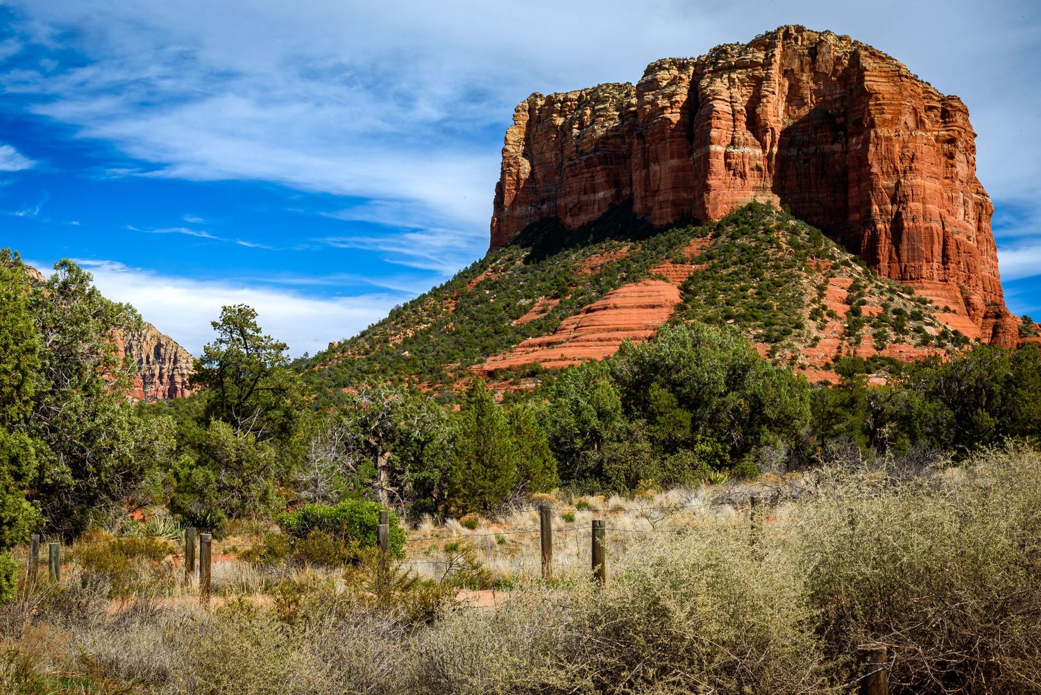 Red rock butte rises above green desert shrubs and trees under blue skies in Sedona, Arizona.