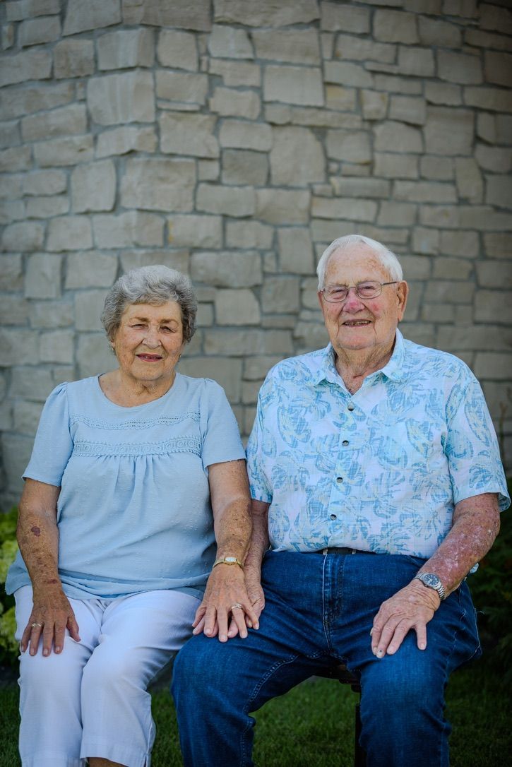 Elderly couple smiling and sitting together outdoors against a stone wall backdrop.