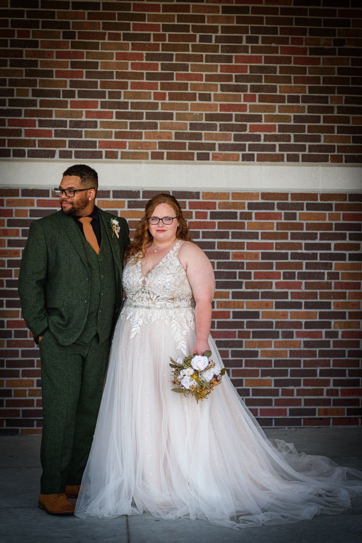 Bride in white tulle gown and groom in green suit pose together against a brick wall on their wedding day.
