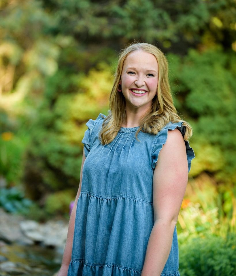 Smiling blonde woman in a blue ruffled dress poses outdoors in a lush green garden setting.