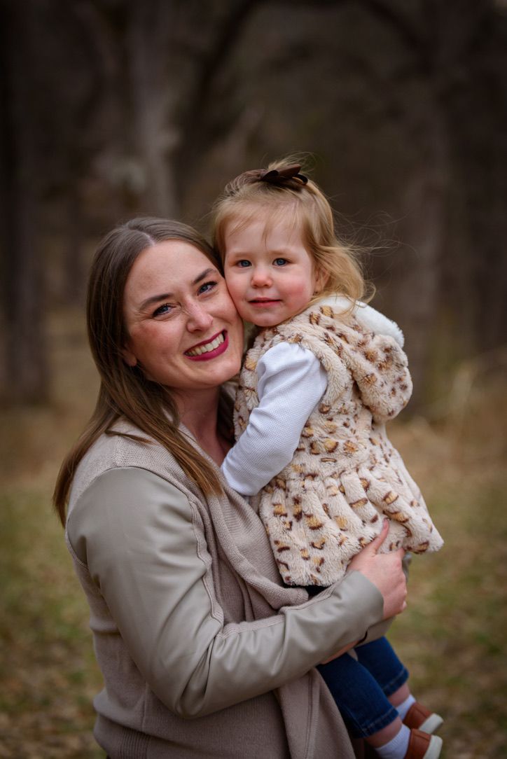 A smiling woman in a beige coat holds a toddler girl in a fuzzy vest outdoors in a wooded setting.