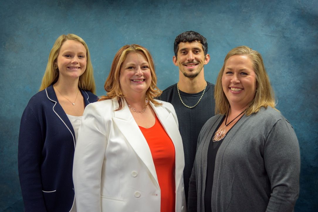 Four professionals smile together for a group portrait against a blue background in business casual attire.