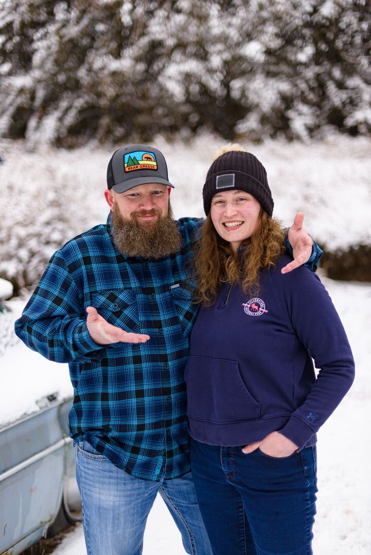 Couple smiling in snowy outdoor setting, man with beard in flannel shirt and woman in navy hoodie and beanie hat.