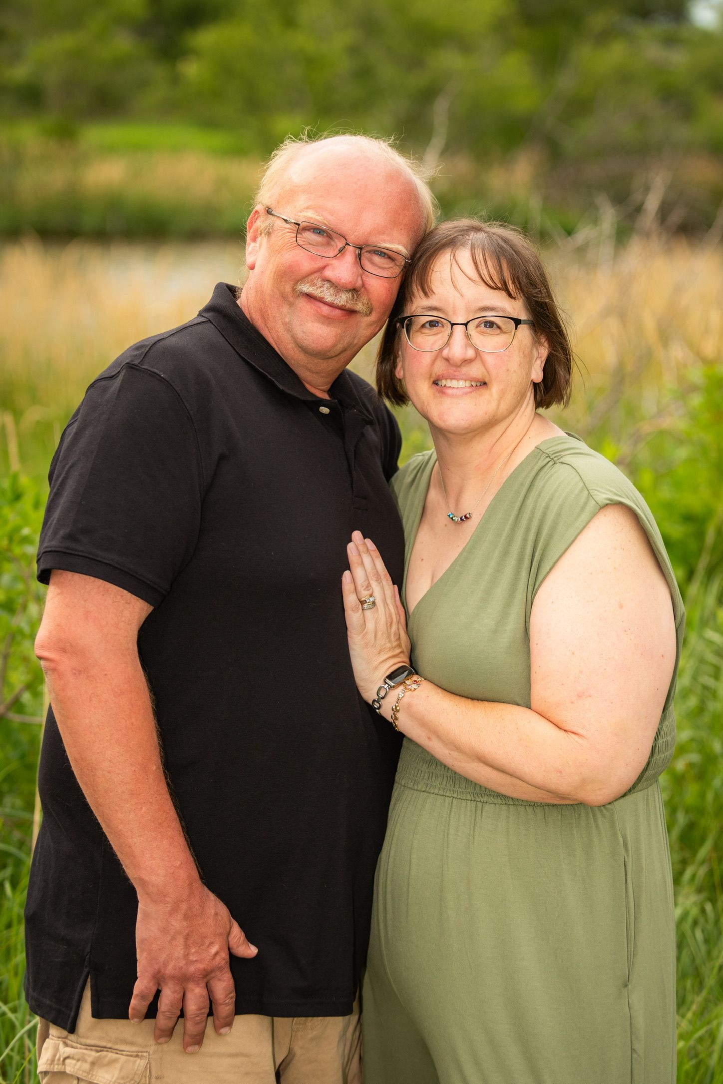A smiling middle-aged couple poses outdoors in a green field, woman in sage dress, man in black polo shirt.