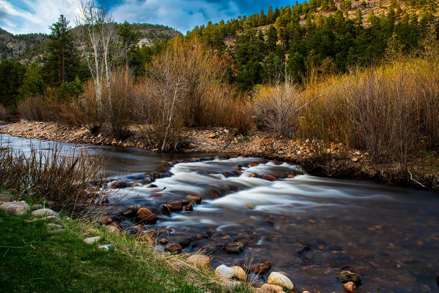A serene mountain stream flows over rocky rapids surrounded by autumn trees and lush greenery in a scenic landscape.