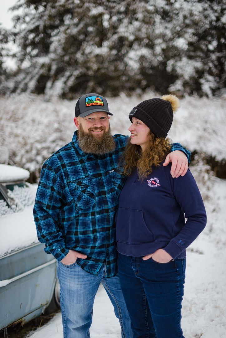 Couple smiling together in snowy winter setting, man in plaid flannel and trucker hat, woman in navy hoodie and beanie.